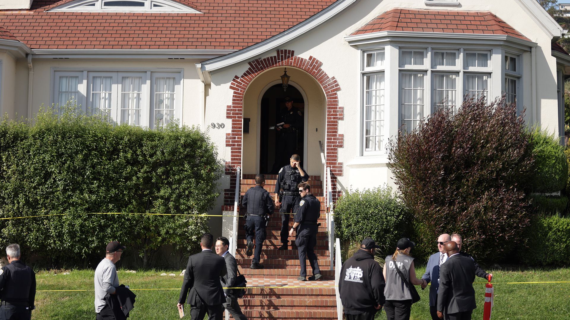 A white, brick-entry house with a curved arched doorway; police officers and detectives stand on red-brick steps, yellow caution tape across the lawn, several suited civilians nearby.