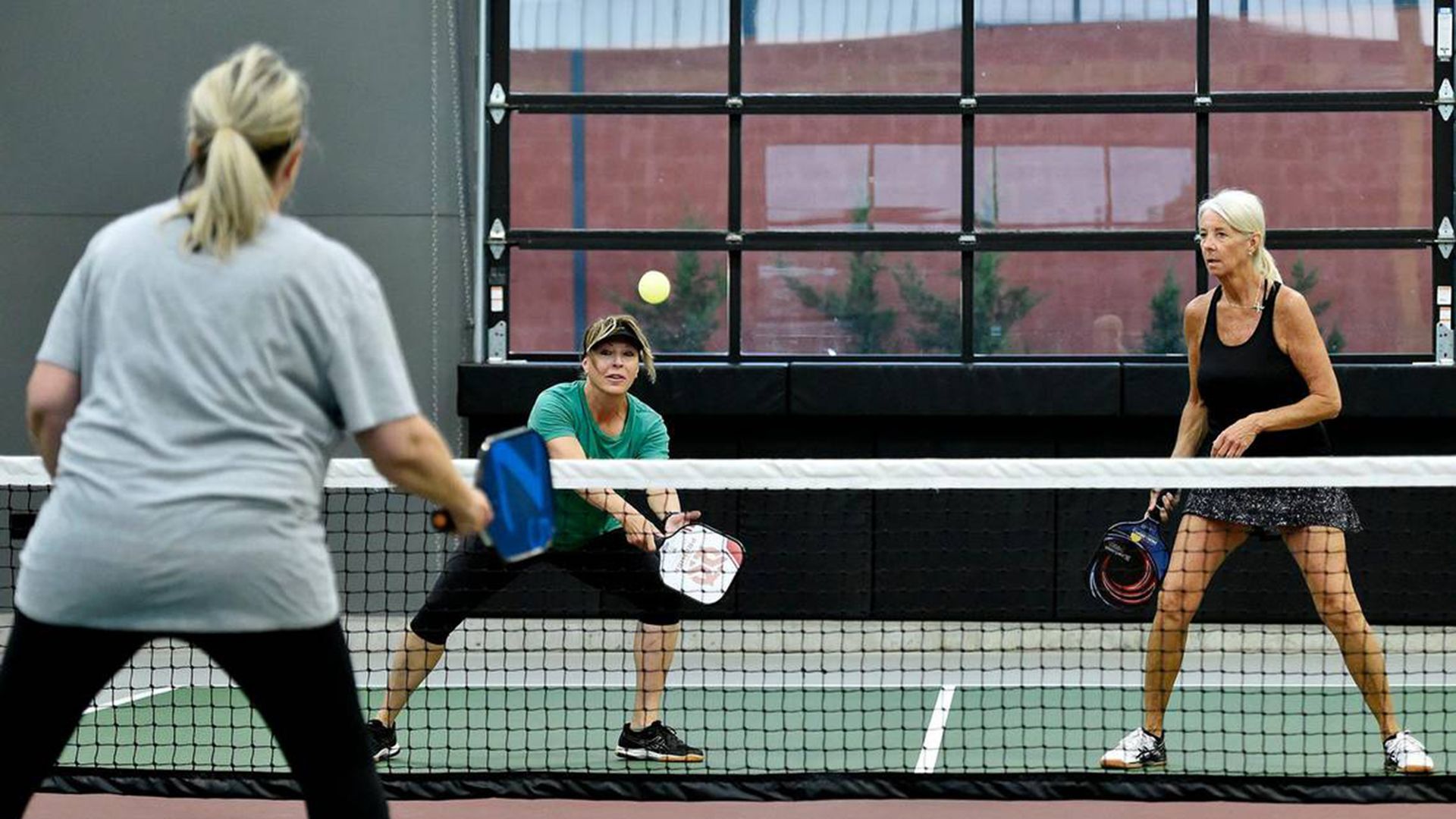 Three women play indoor pickleball at a net: a blonde in gray on the left, a green-shirted croucher in the middle, and a woman in black on the right, chasing a yellow ball near a brick wall.