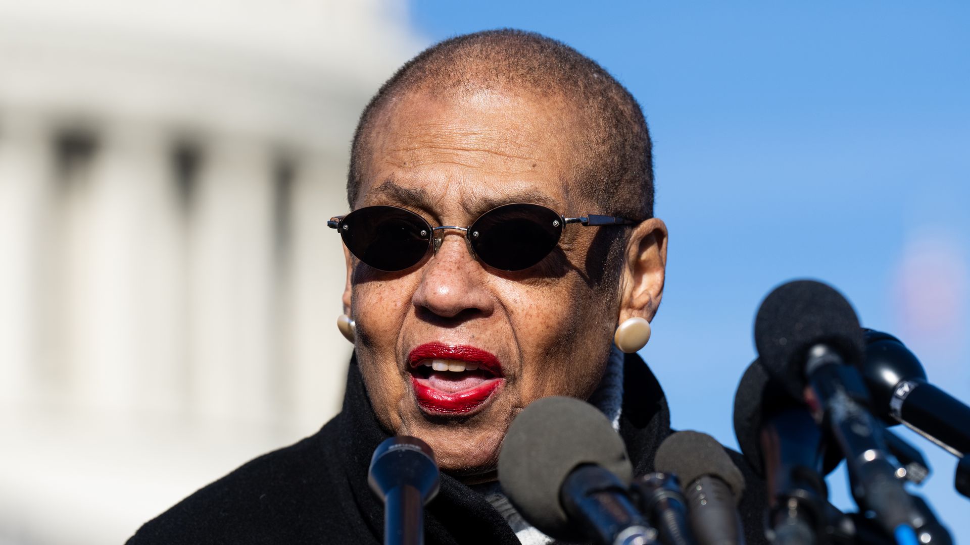Rep. Eleanor Holmes Norton, wearing black circular sunglasses and speaking at microphones in front of the Capitol dome.