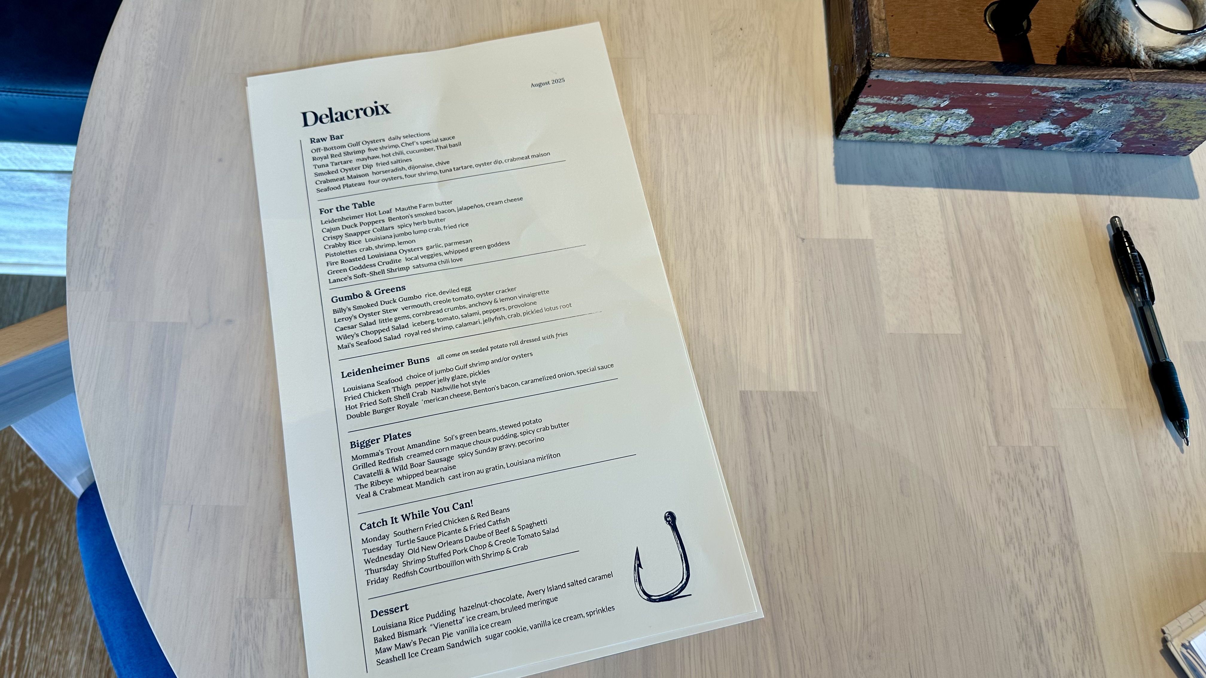 Menu titled Delacroix dated August 2025 on a light wood table with dishes categorized under Raw Bar, For the Table, Gumbo & Greens, Leidenheimer Buns, Bigger Plates, Catch It While You Can!, and Dessert, with a fishing hook illustration.