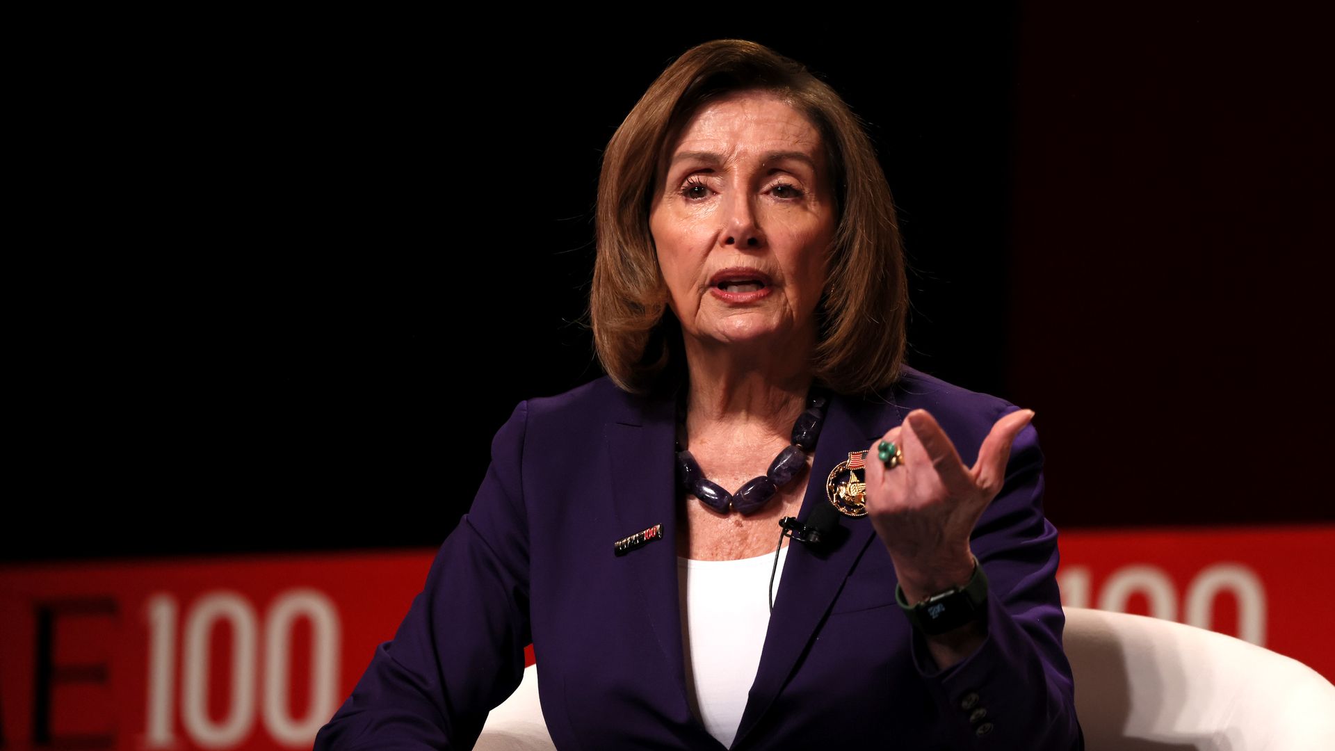 Former House Speaker Nancy Pelosi wearing a purple suit and sitting in front of a black wall.