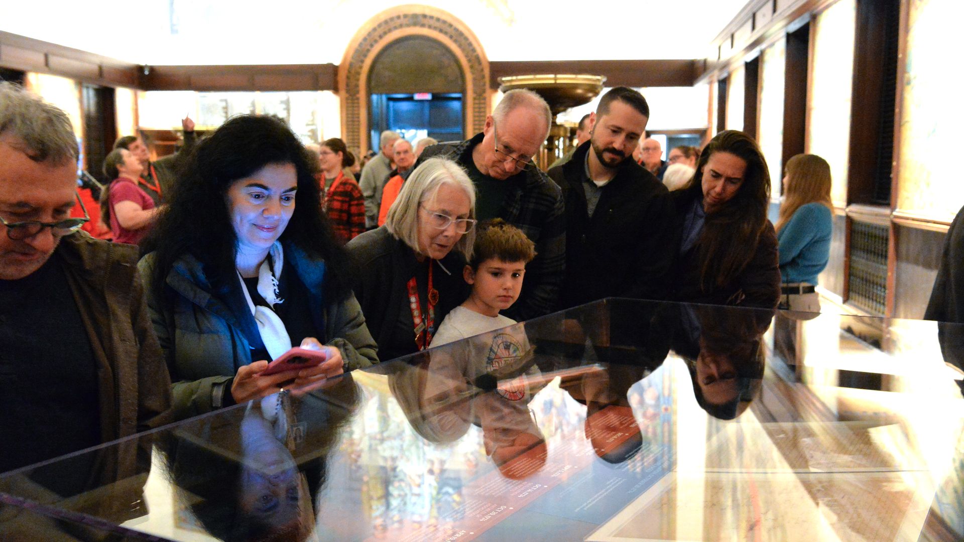 A group of museum visitors leans over a glass display case, studying illuminated panels. A woman with dark hair uses a smartphone while a child and several adults observe in a bright, ornate hall.