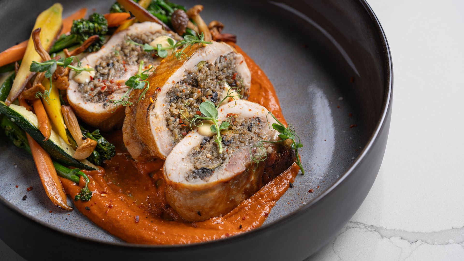 Three slices of stuffed chicken on red sauce with assorted sautéed vegetables including carrots, zucchini, and mushrooms in a black bowl on a white marble surface.