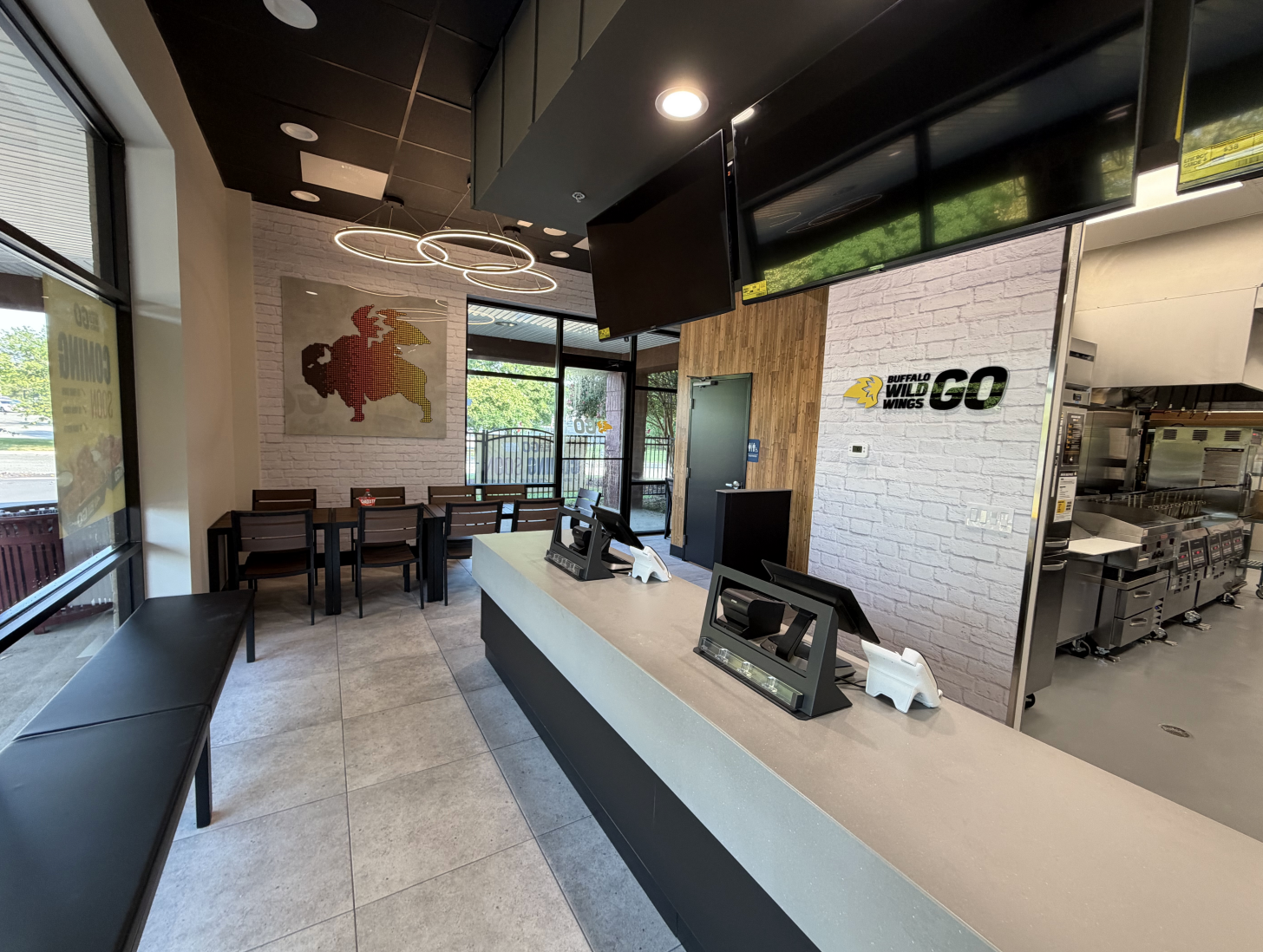 Modern fast-casual interior with a long white counter, black trim, circular ring lights, white brick walls, and a Buffalo Wild Wings GO sign. Tables and chairs by large windows; kitchen area visible.