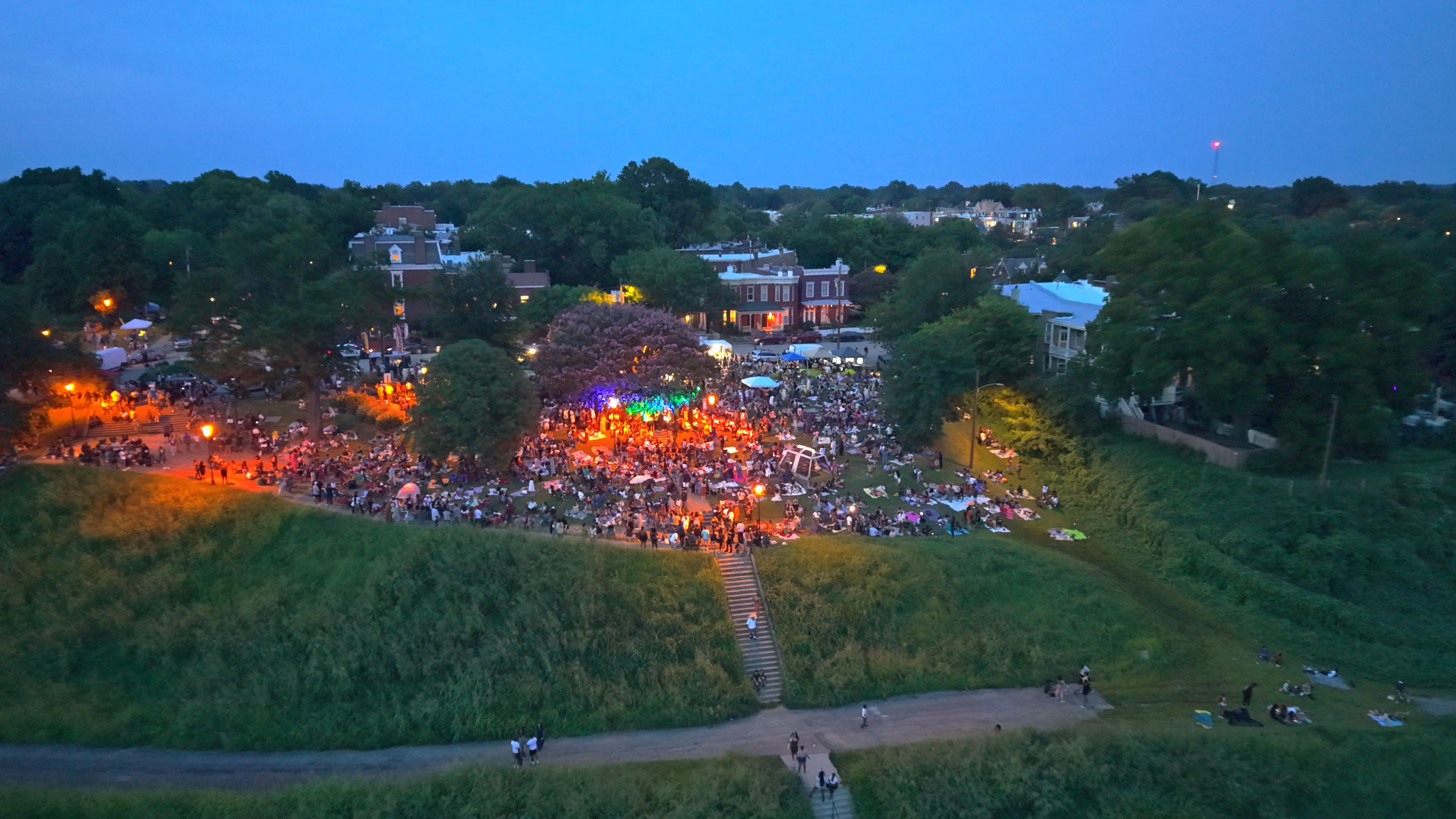 An aerial dusk view of a crowded park festival: a lit stage with colorful lights, tents, and many people gathered, surrounded by trees and houses in the background.