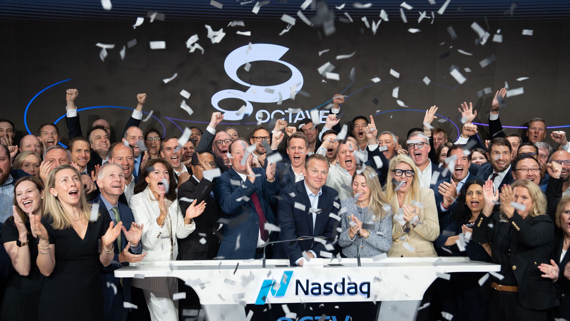 A large crowd of suited professionals on a Nasdaq stage celebrate with raised fists and confetti, behind them a prominent Nasdaq logo backdrop.