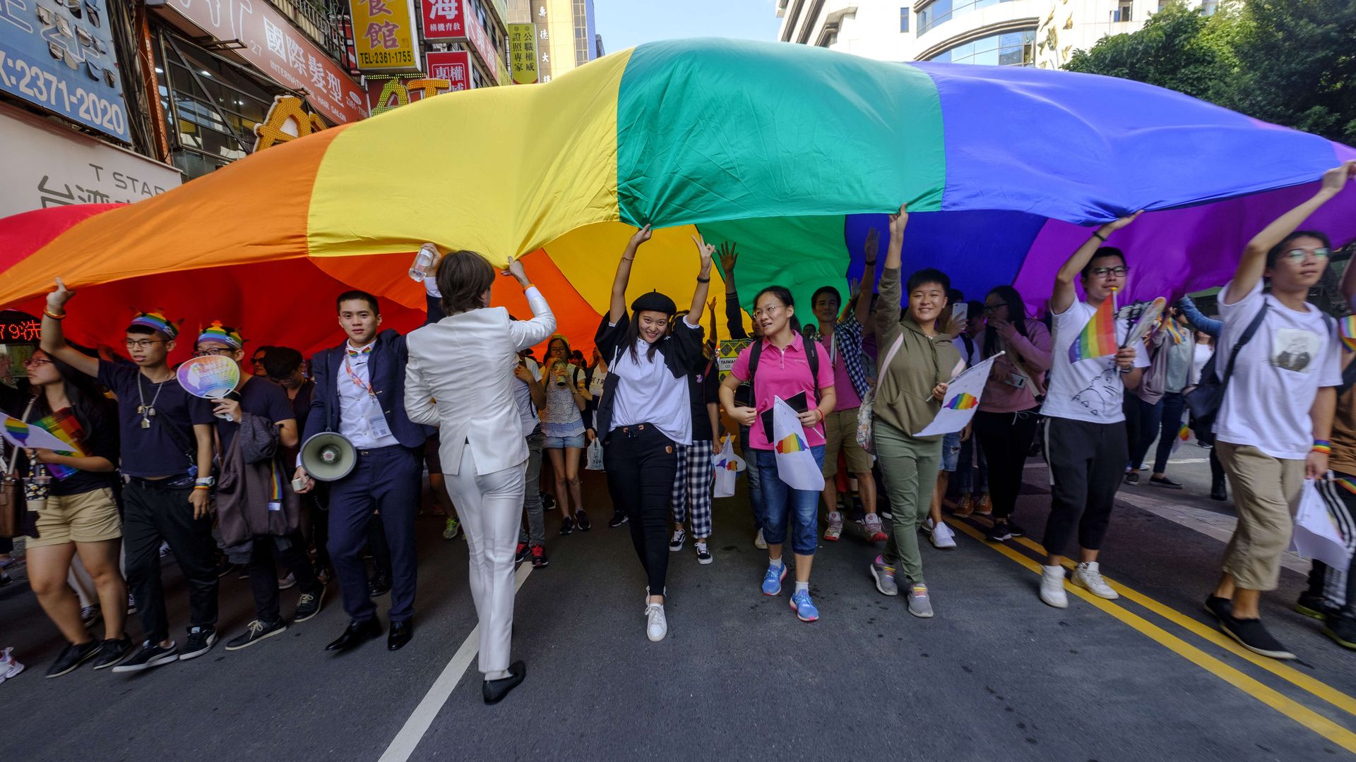 In this image, a line of demonstrators walk down the street carrying a large rainbow flag aloft.