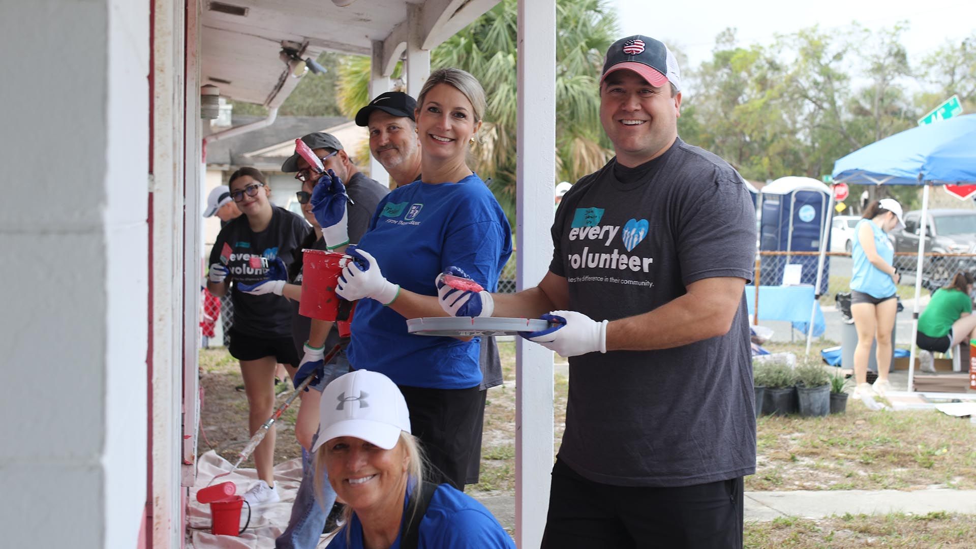 Group of smiling volunteers wearing gloves and casual clothes, painting a house exterior pink outdoors with trees and tents in the background on a cloudy day.