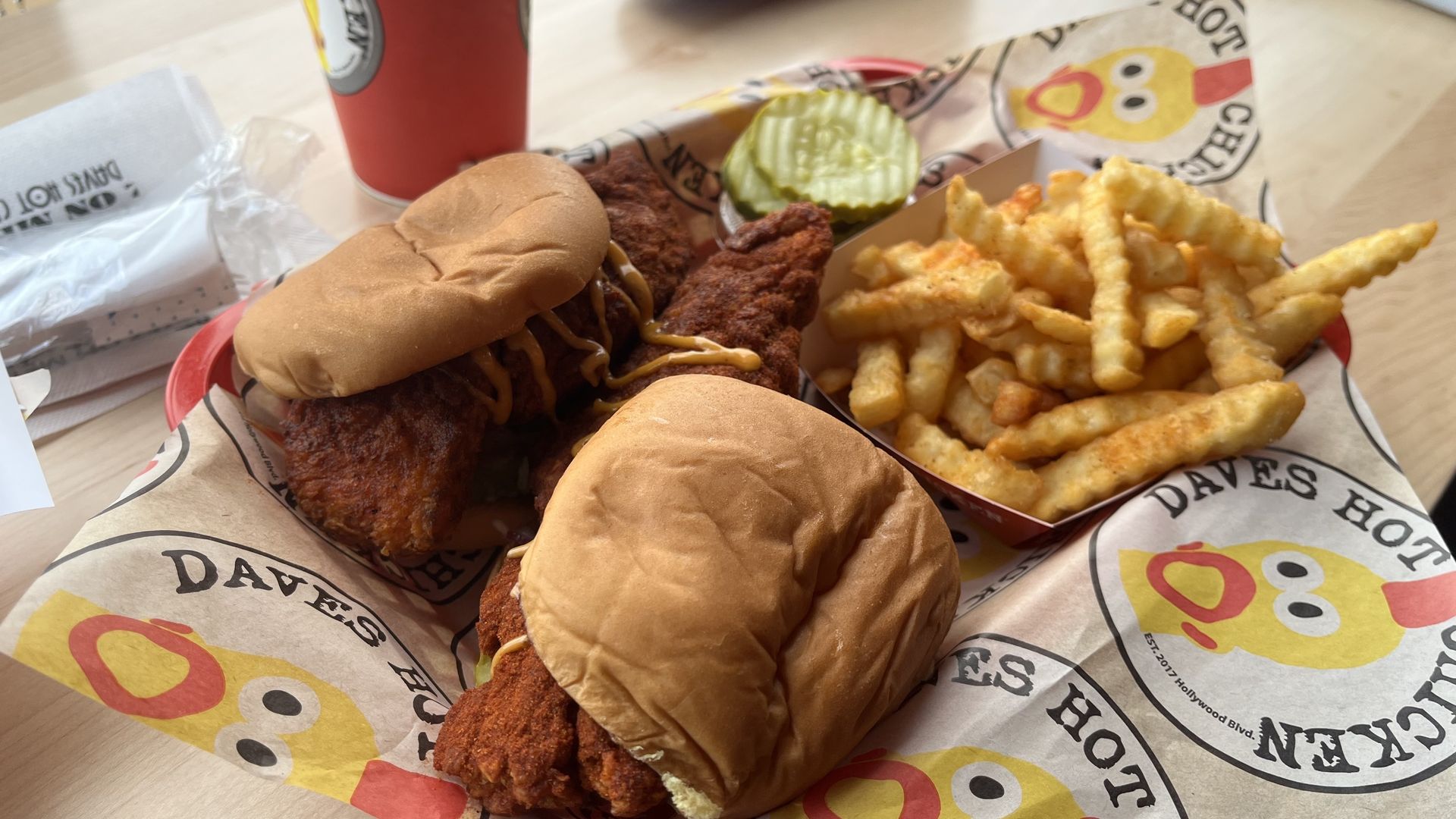 Two fried chicken sliders with drizzle on buns, a heap of crinkle-cut fries, a pickle slice, and a red soda cup on a table; Dave's Hot Chicken wrappers visible in the background.