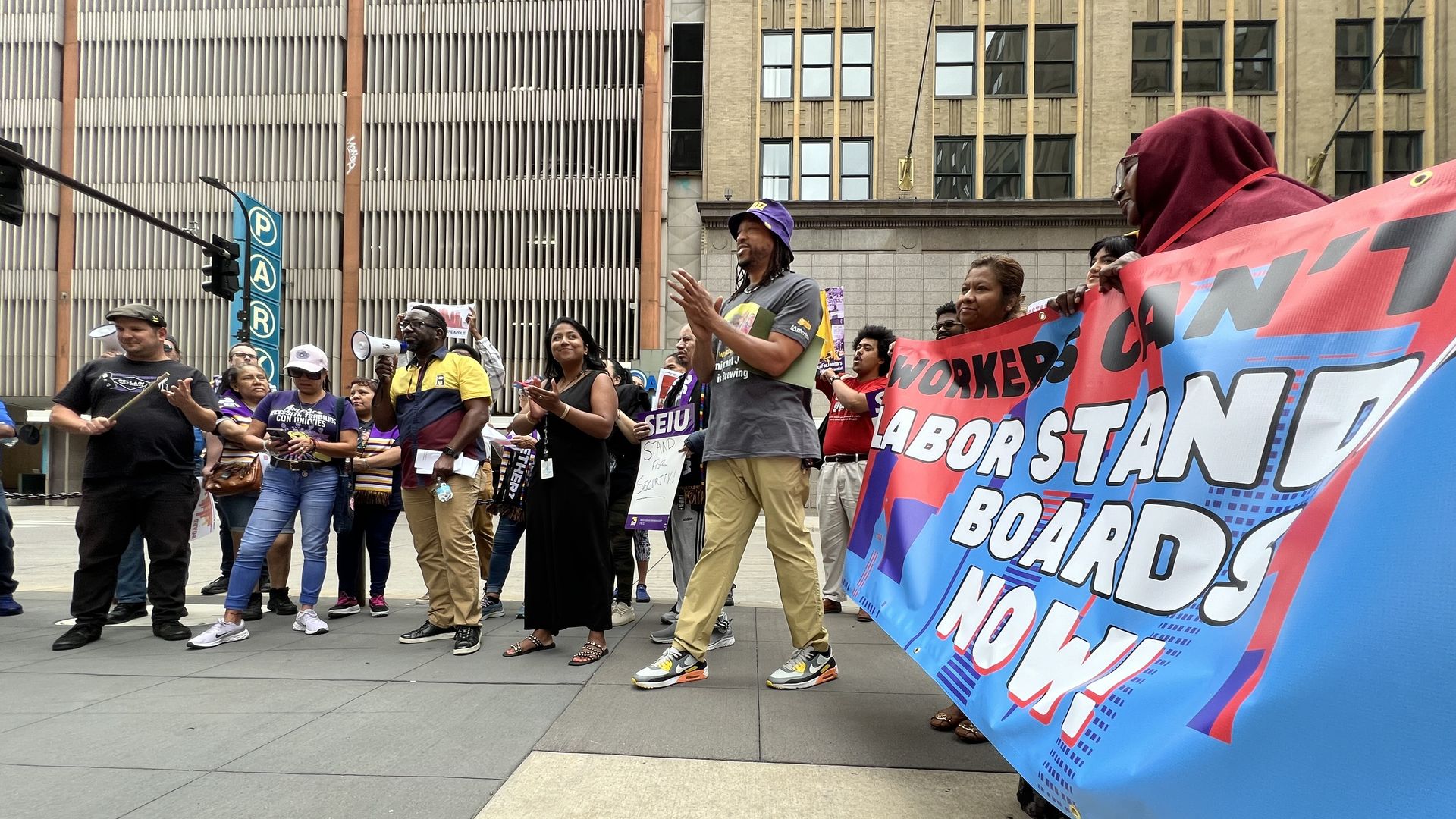 Rally-goers hold a large banner reading "Labor Standards Boards Now!" at an outdoor demonstration on a downtown corner