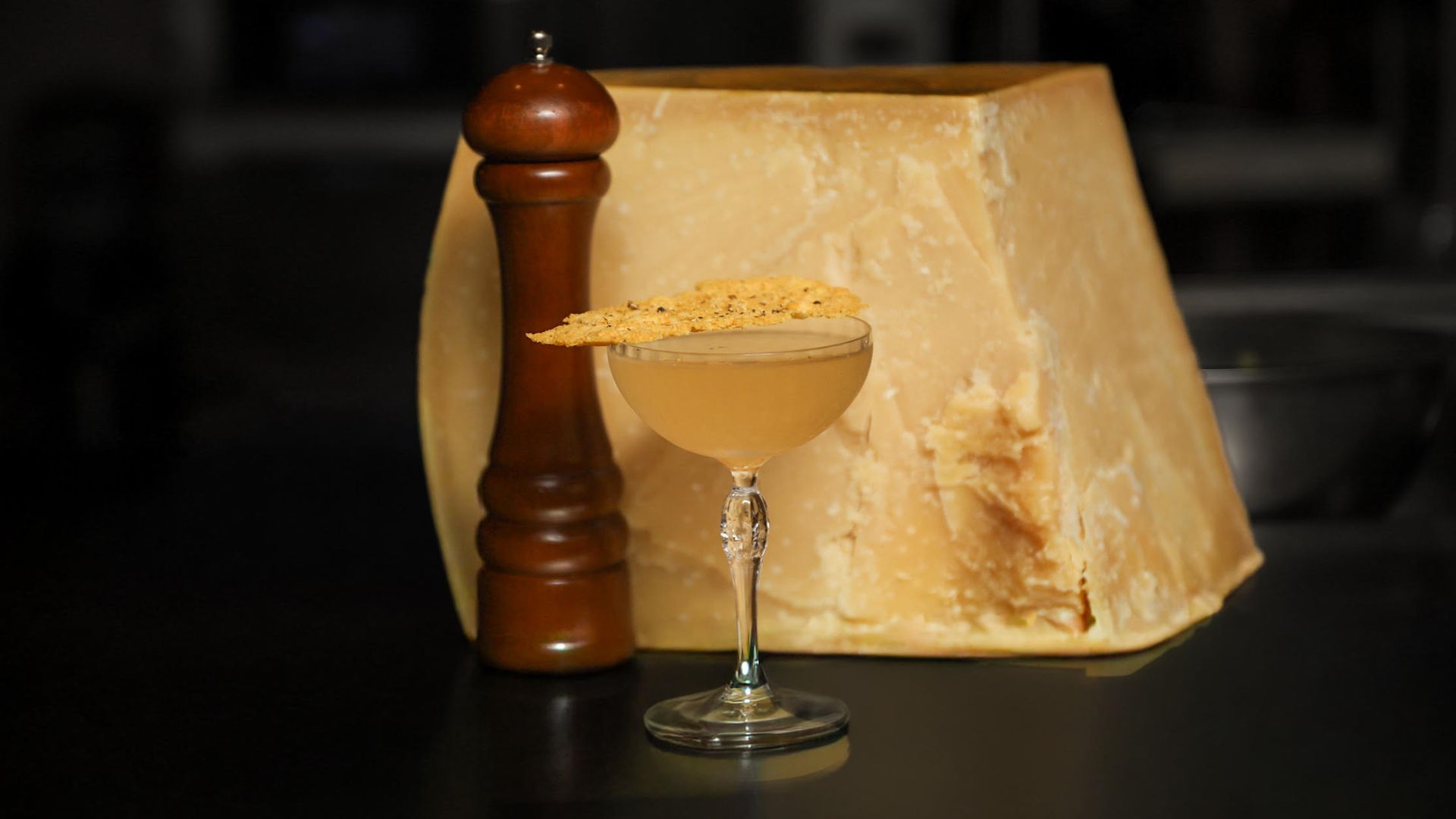A large wedge of pale yellow cheese, a wooden pepper mill, and a cocktail in a coupe glass garnished with a thin, crispy cracker on top, all set on a dark surface.