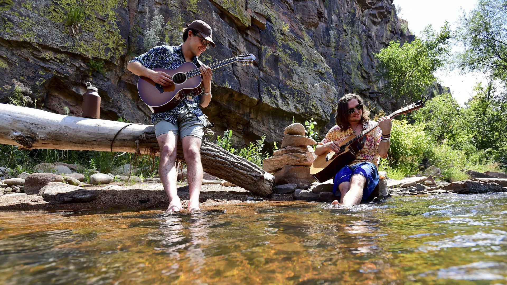 Two young men playing acoustic guitars by a clear stream, one seated in the water and one standing on the shore, surrounded by rocky cliffs and green trees.