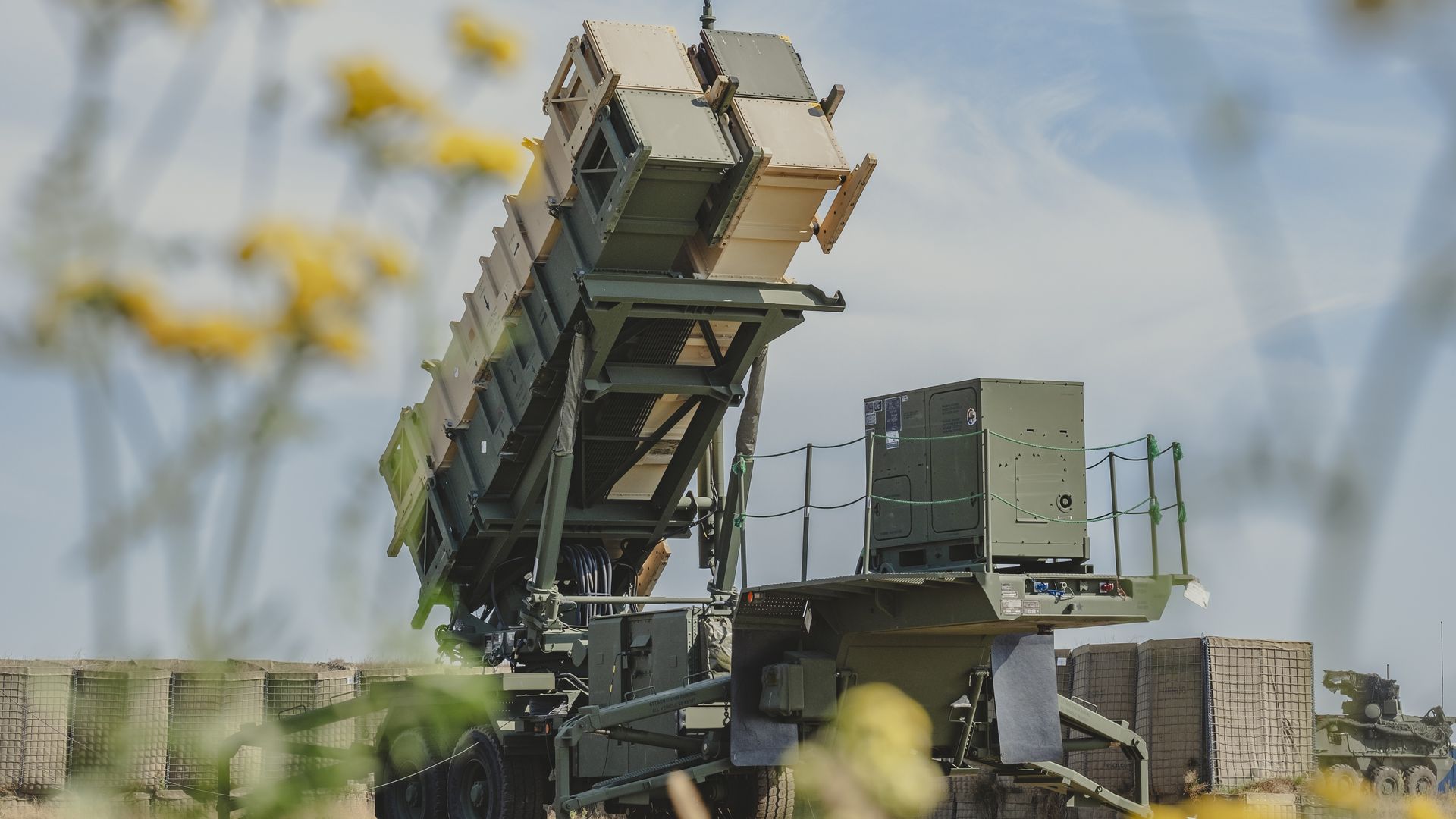 Military missile launcher system in green and khaki, angled upward, surrounded by yellow wildflowers with a blue sky and protective barriers in the background.
