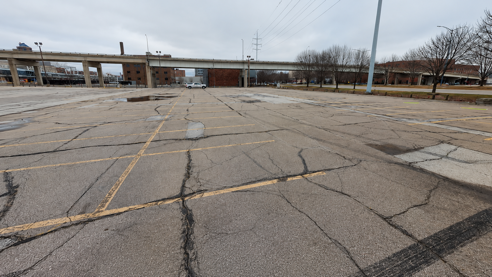Wide, cracked asphalt parking lot with faded yellow lines and puddles, empty space toward a distant white car under an overpass, bare trees and gray, cloudy sky.