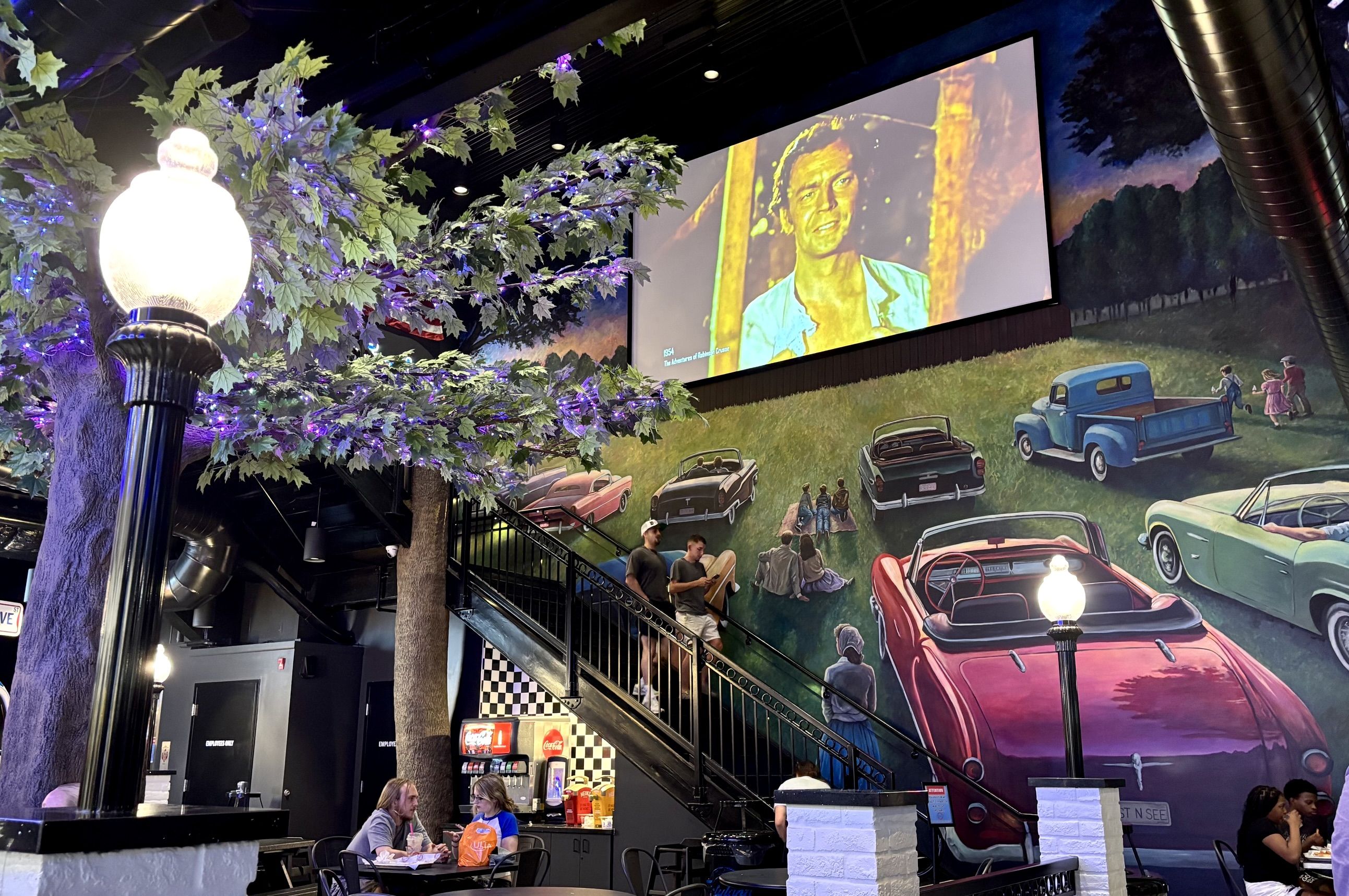 Indoor space with a mural of colorful vintage cars, a glowing lamp post among leafy trees with purple lights, and a large screen showing a portrait; people dine at tables.
