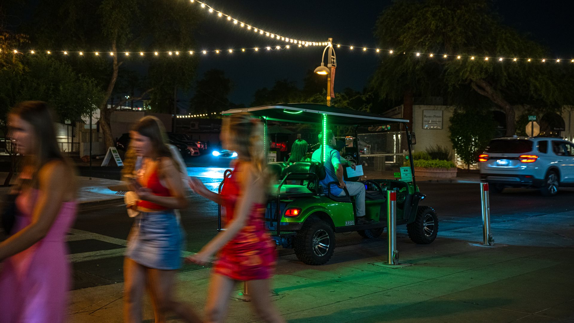 Three girls walking, gold cart in the background and string lights overhead and brightly lit in the dark sky.