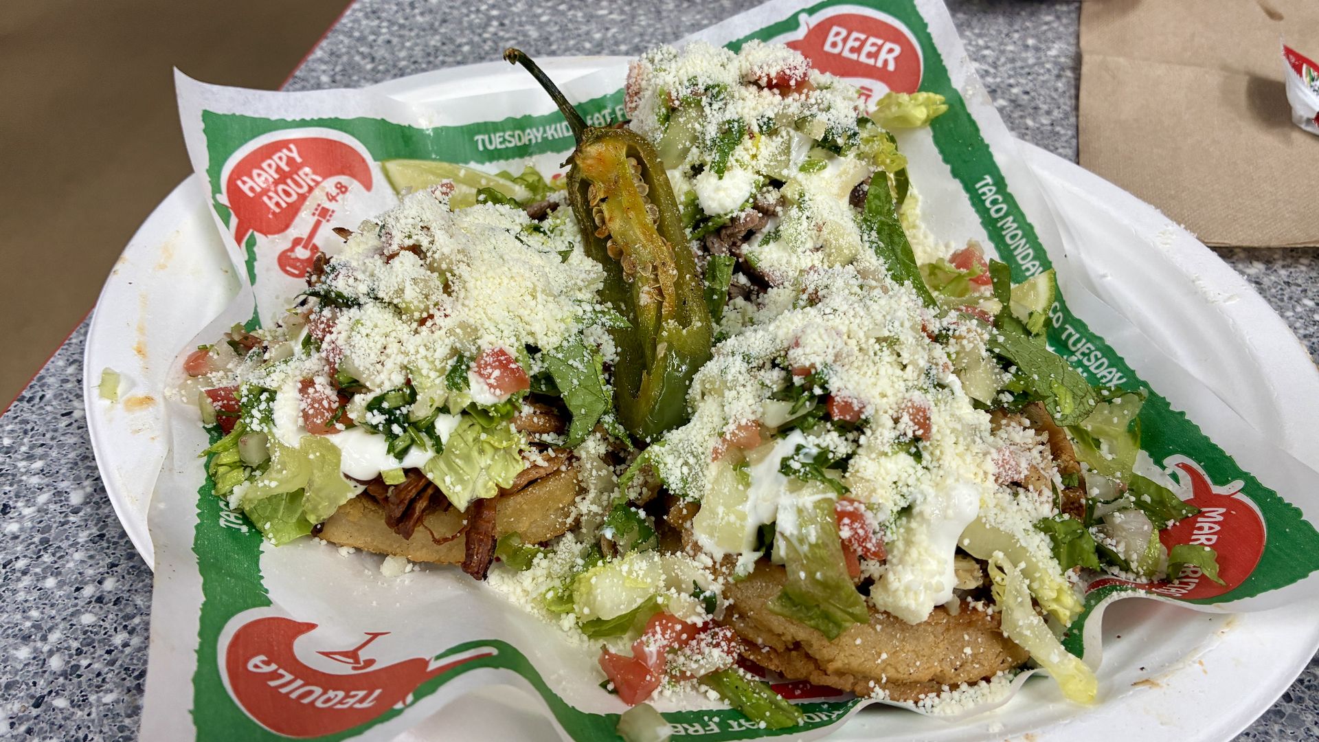 A loaded street-style sopes on a plate with a green wrapper, topped with cotija cheese, lettuce, diced tomatoes, cilantro, crema, and a jalapeño standing upright.