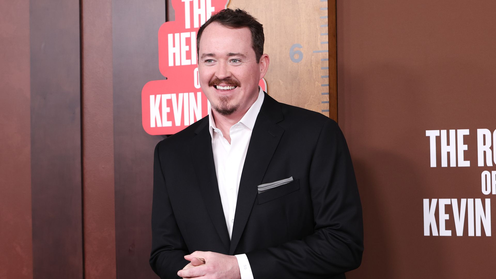 Smiling man in a black suit and white shirt with a patterned pocket square, hands clasped, stands before a brown backdrop with large white lettering and the words "THE KEVIN" behind him.