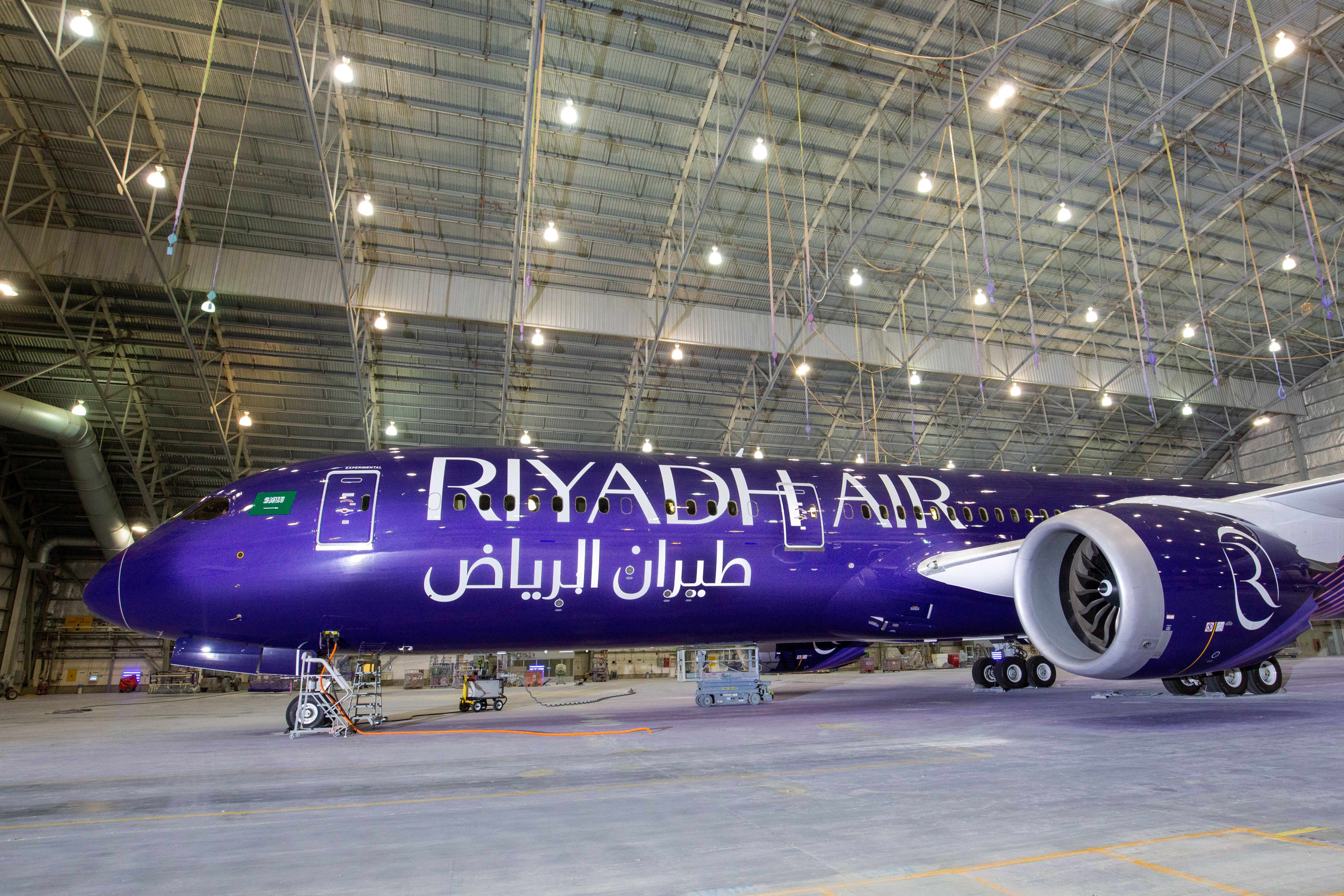 Purple Riyadh Air airplane inside a large hangar with bright overhead lights, displaying the airline's name in English and Arabic on the fuselage.