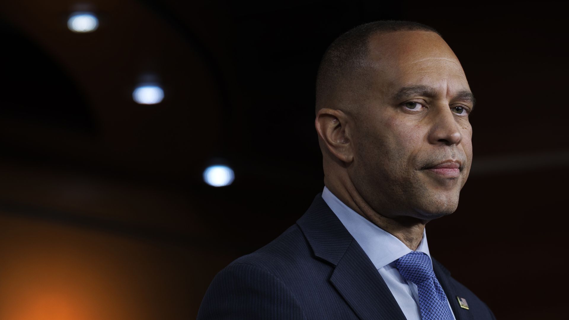 Hakeem Jeffries, wearing a blue suit and speaking in front of an orange wall below a handful of lights on a dark ceiling.