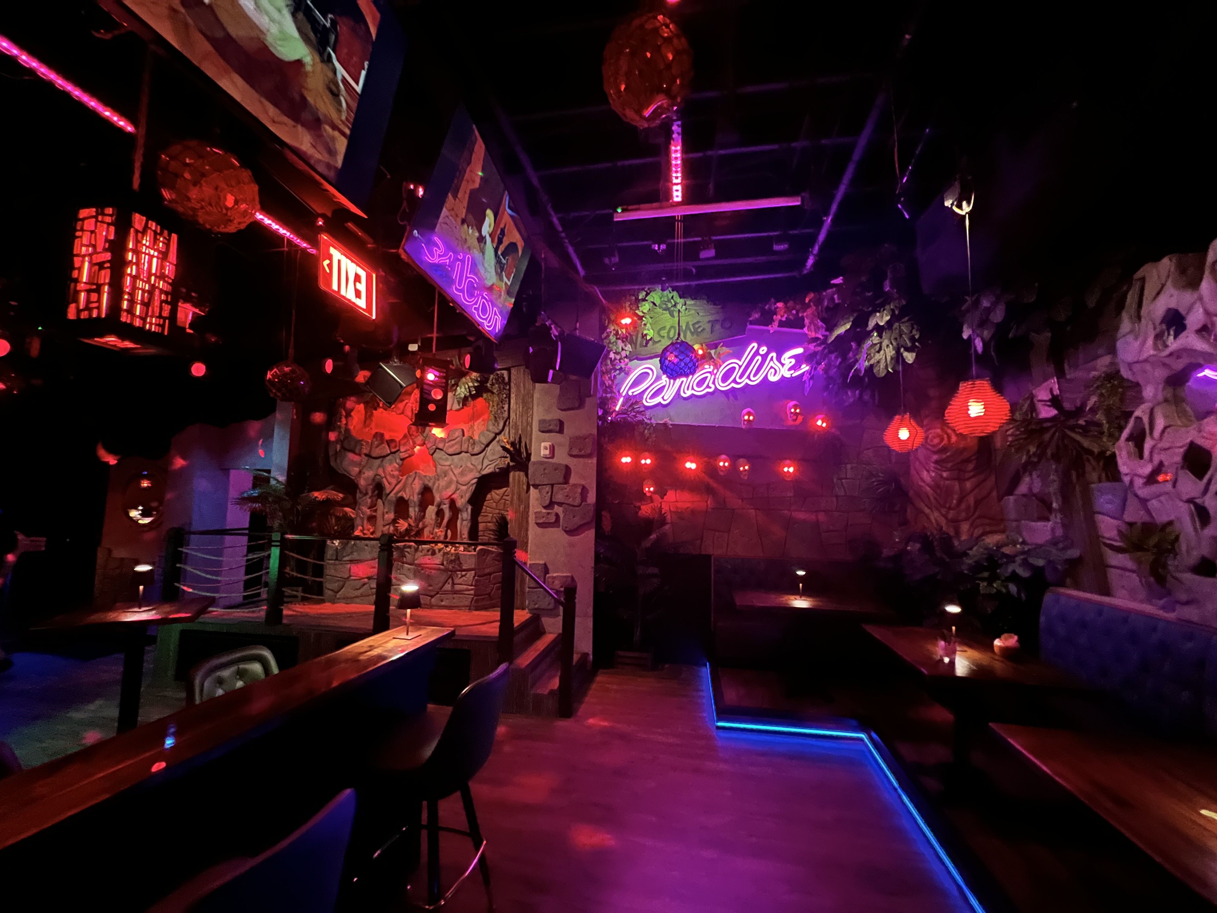 Nightclub interior with neon "Paradise" sign, pink/red lighting, tropical plants, rock walls, booths along a blue LED floor edge; hanging red lanterns and an exit sign in view.