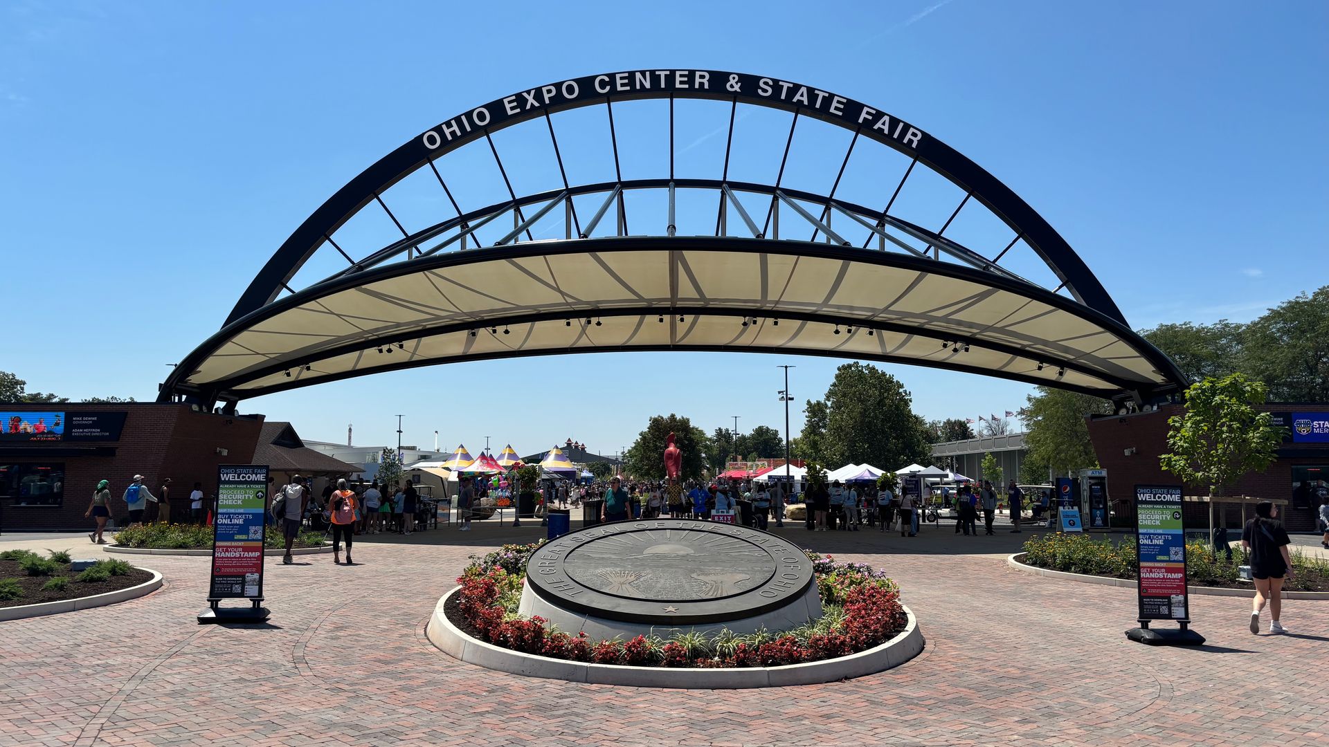 Wide view of the Ohio Expo Center & State Fair arch under a clear blue sky. A large black curved arch with white lettering spans a circular plaza, with tents, people, and brick-paved walkways around it.