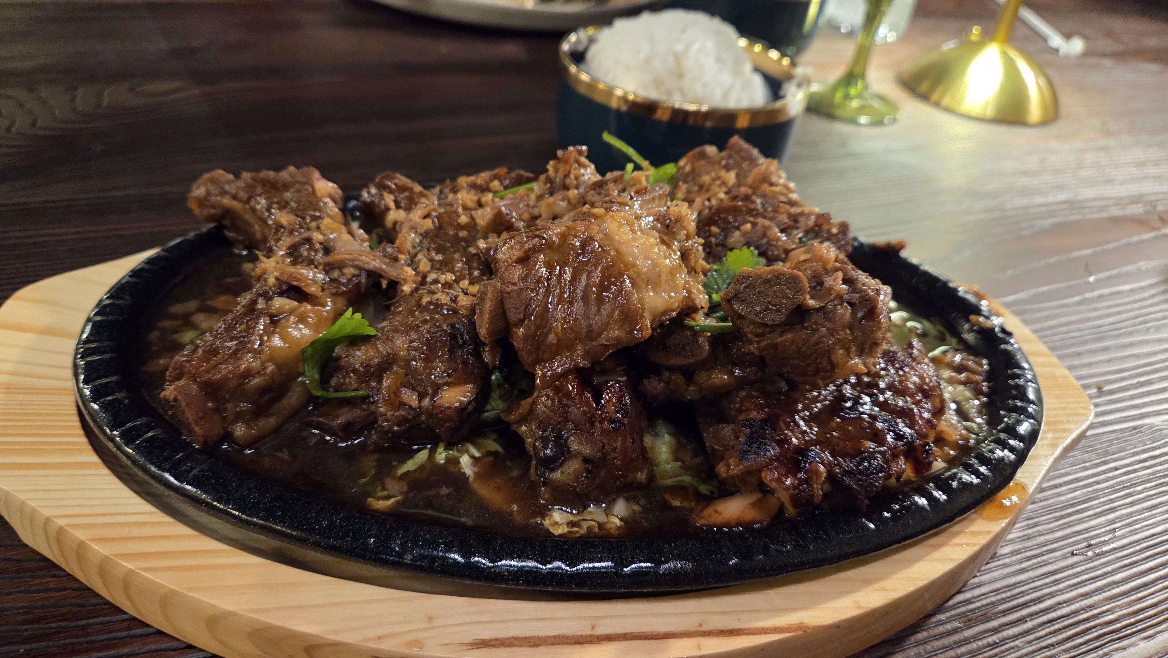Plate of braised pork ribs in dark sauce garnished with cilantro, served on a black sizzling platter with wooden base, with a bowl of white rice and drinks in the background.