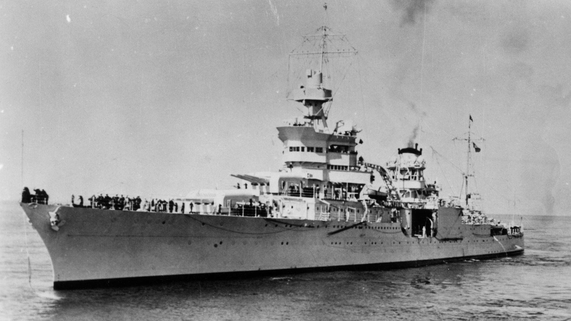 Black and white photo of a large naval warship with guns and antennas, sailing on calm water with crew visible on deck, emitting smoke from the rear chimney.