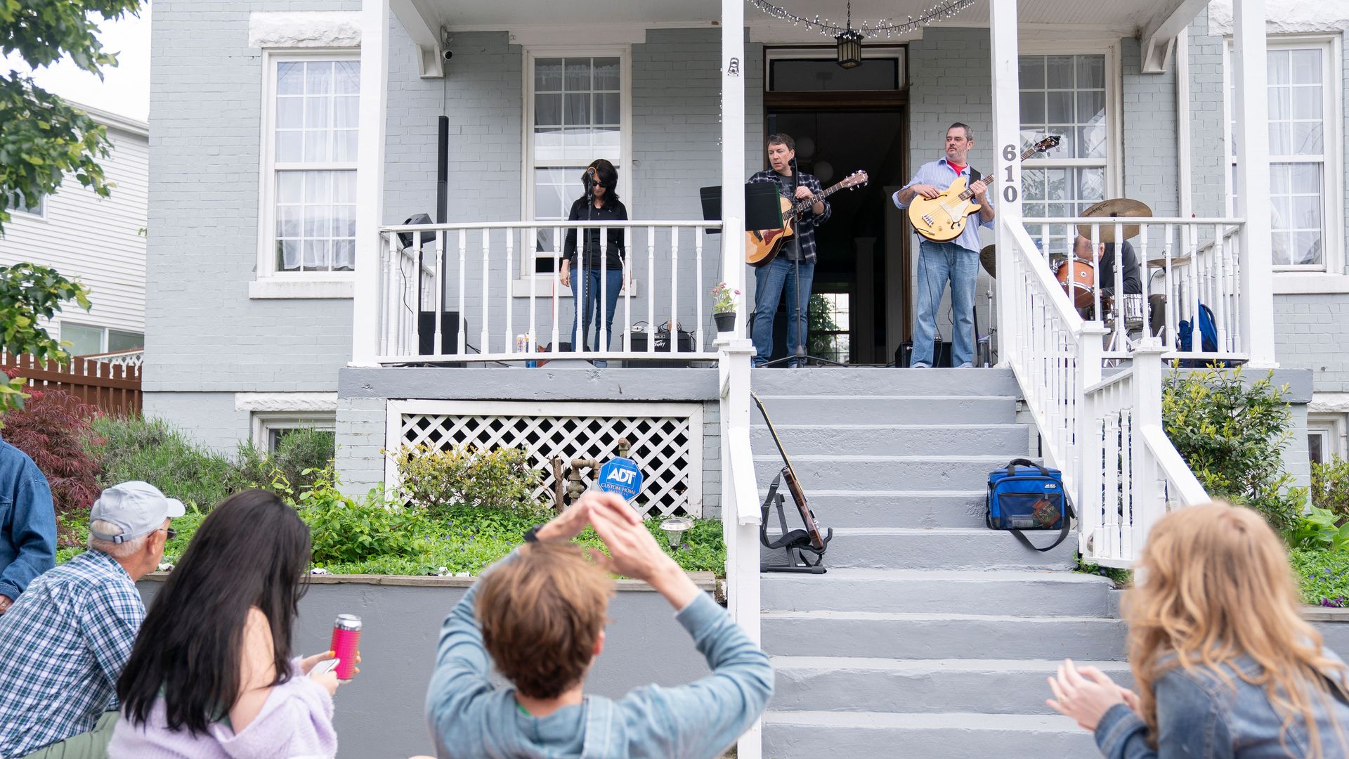 A live outdoor band performs on a gray porch with white railings. Three musicians play guitars and drums while a crowd sits and claps; string lights hang above.