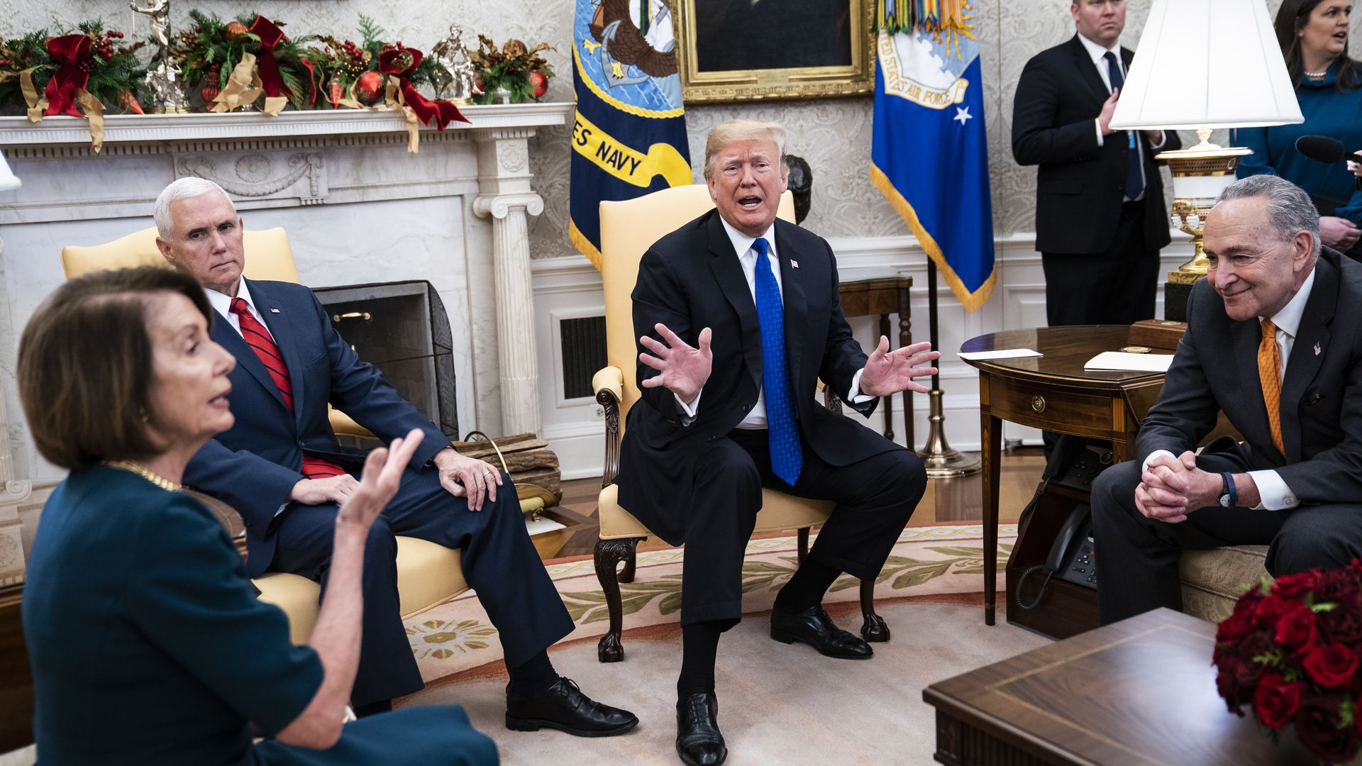 Presidnt Trump waves hand, Nancy Pelosi raises one hand, and Chuck Schumer sits clasping hands.