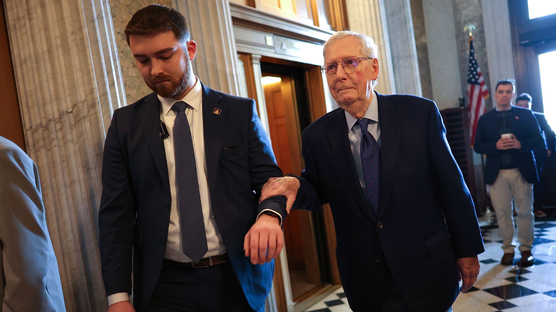 A man in a navy suit and glasses holding the arm of another man in a dark suit and tie, walking indoors by marble columns with people and an American flag in the background.