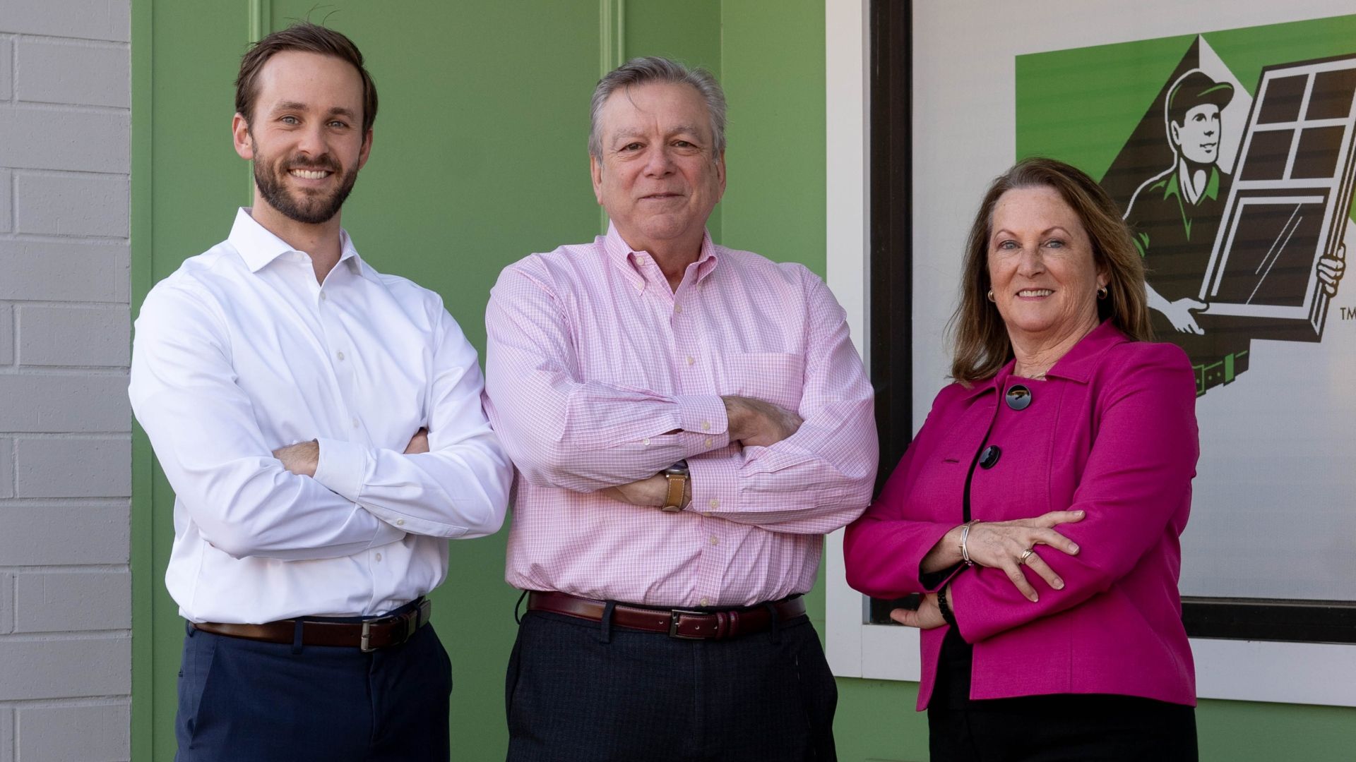 Three adults stand with arms crossed in front of a green wall: left man in white shirt, center older man in pink checkered shirt, right woman in bright pink blazer; all smiling.
