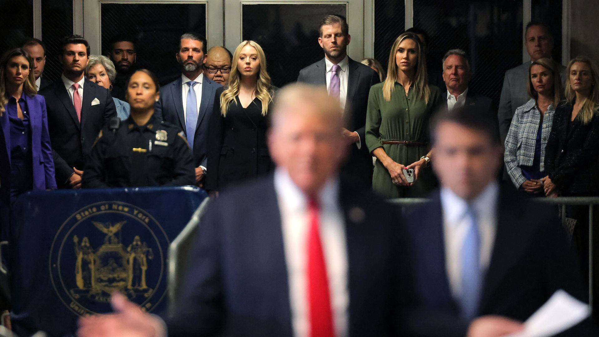 As Donald Trump speaks to reporters (foreground), Trump family members (background) listen. 