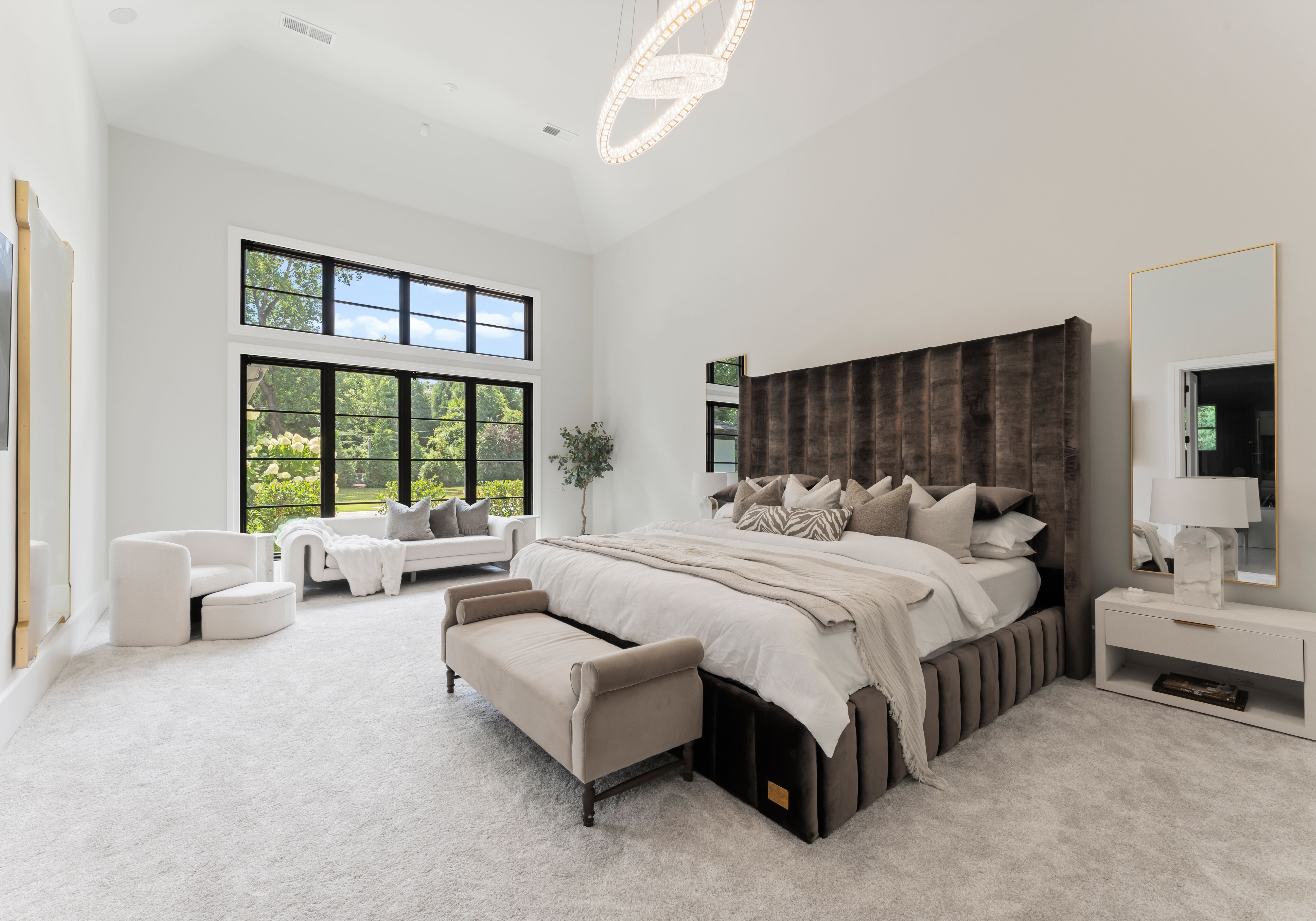 Bright bedroom with large windows, white walls, gray carpet, dark tufted bed, beige bench, white sofa with gray pillows, a cozy armchair, a tall mirror, and modern chandelier.
