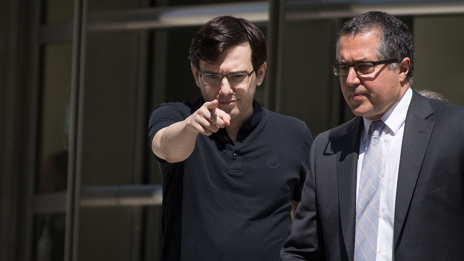 In this image, Martin Shkreli wears a black polo shirt and points at the camera. 