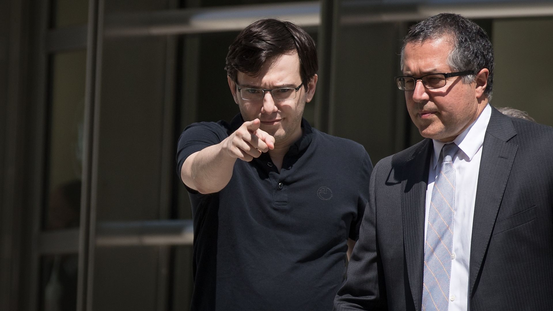 In this image, Martin Shkreli wears a black polo shirt and points at the camera.