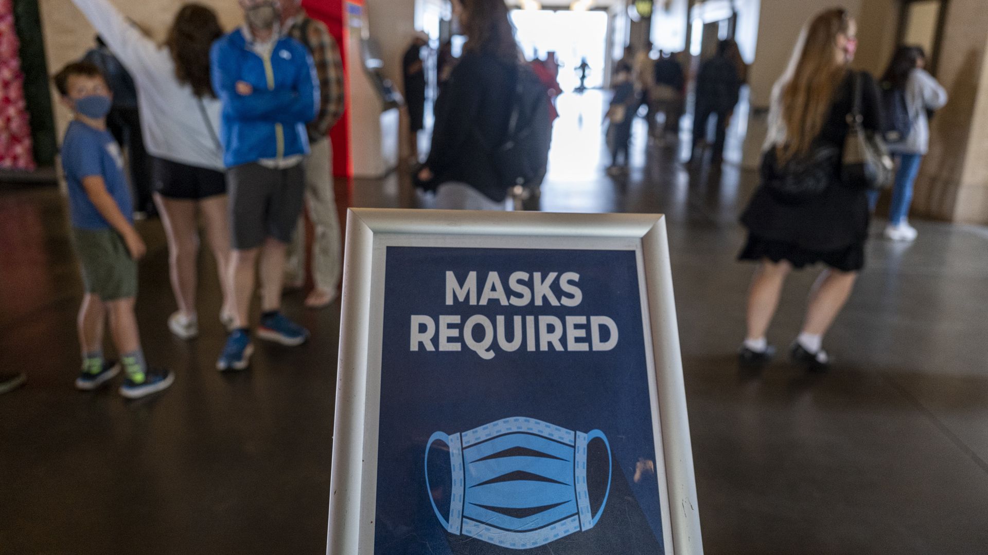 Photo of a sign that says "masks required" as people walk around in the background