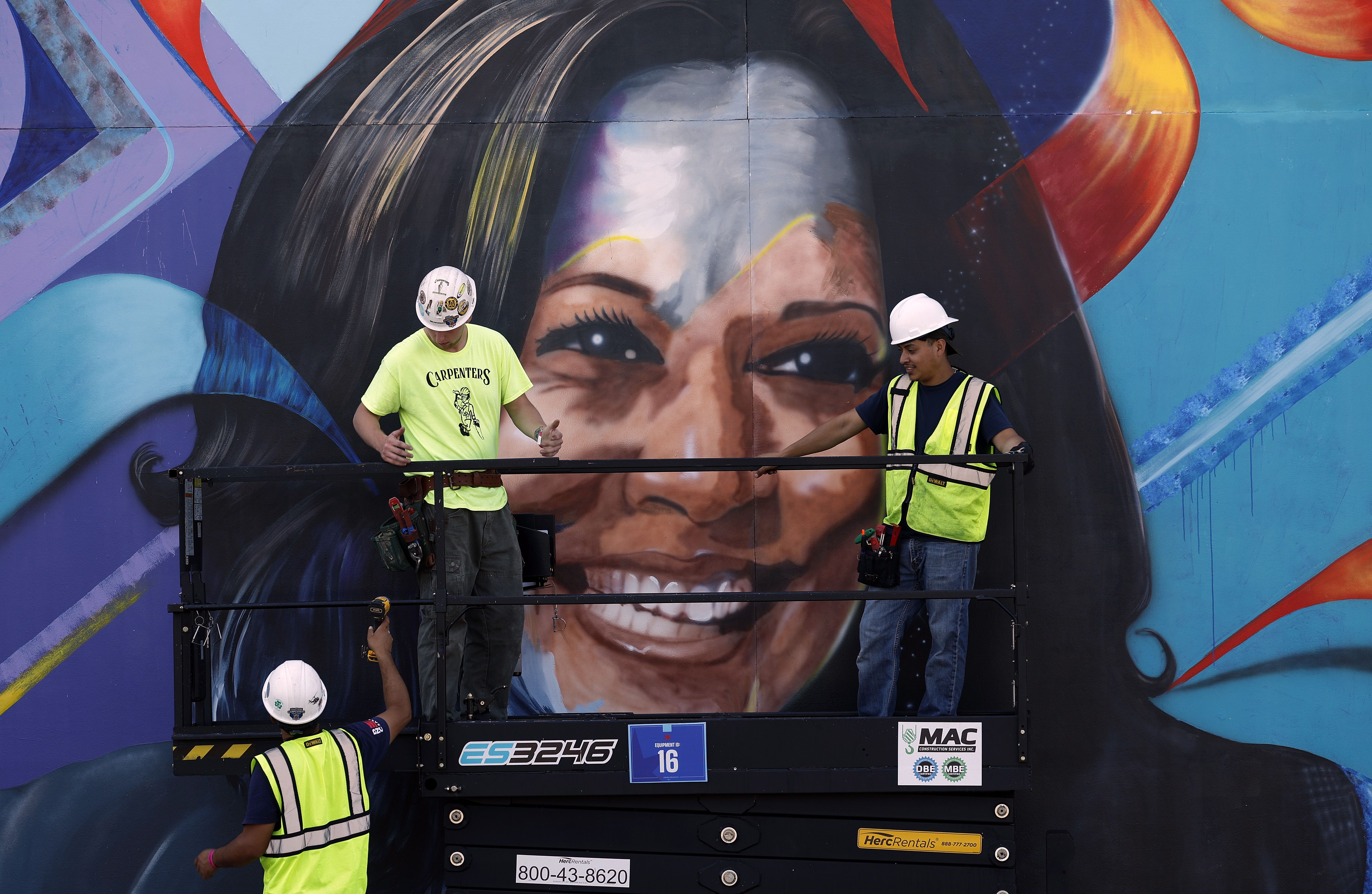 Three people in hard hats work on a large mural