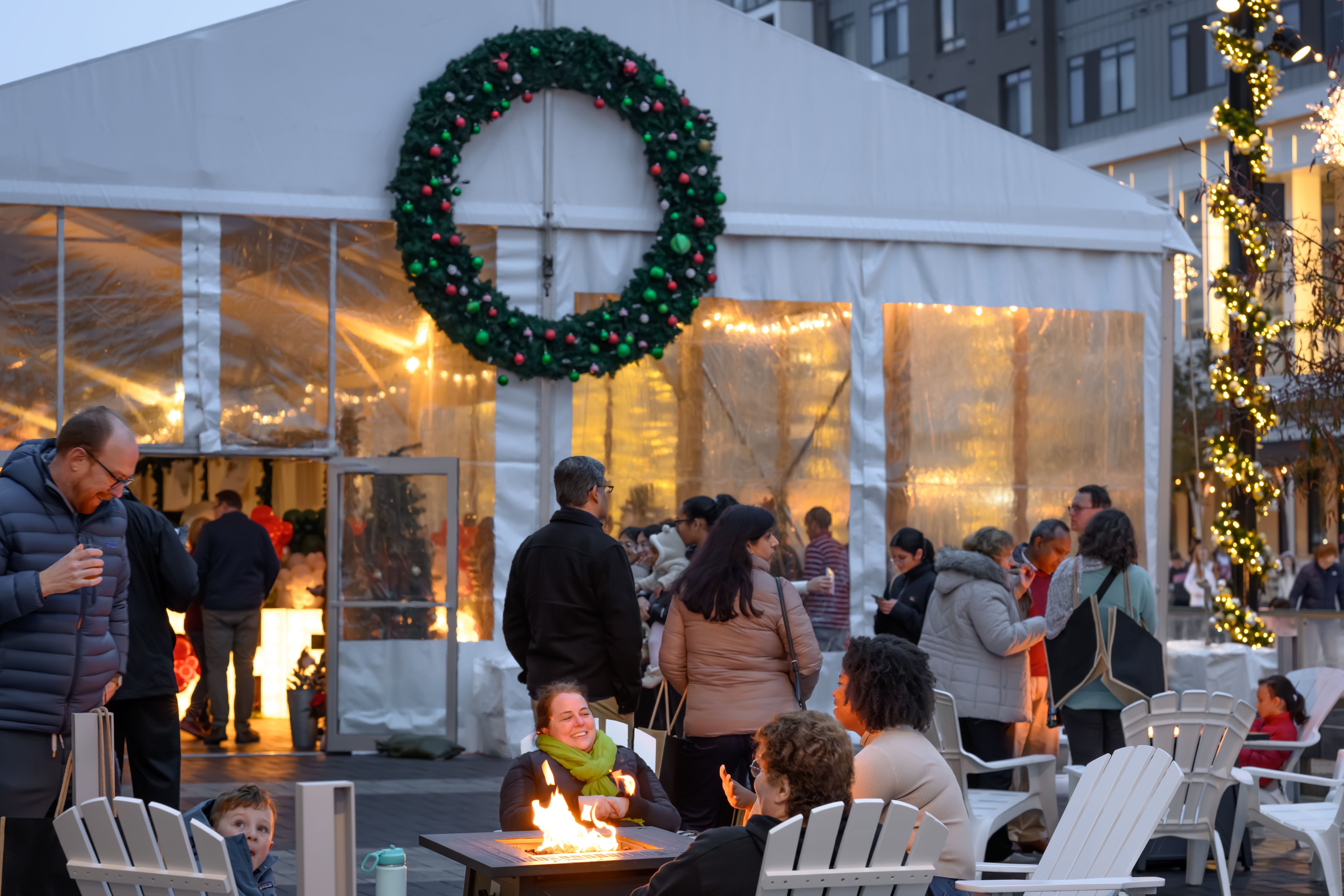 People gathered around a fire pit outside a white tent decorated with a large green wreath with red and green ornaments; festive lights wrapped on poles create a warm holiday atmosphere.