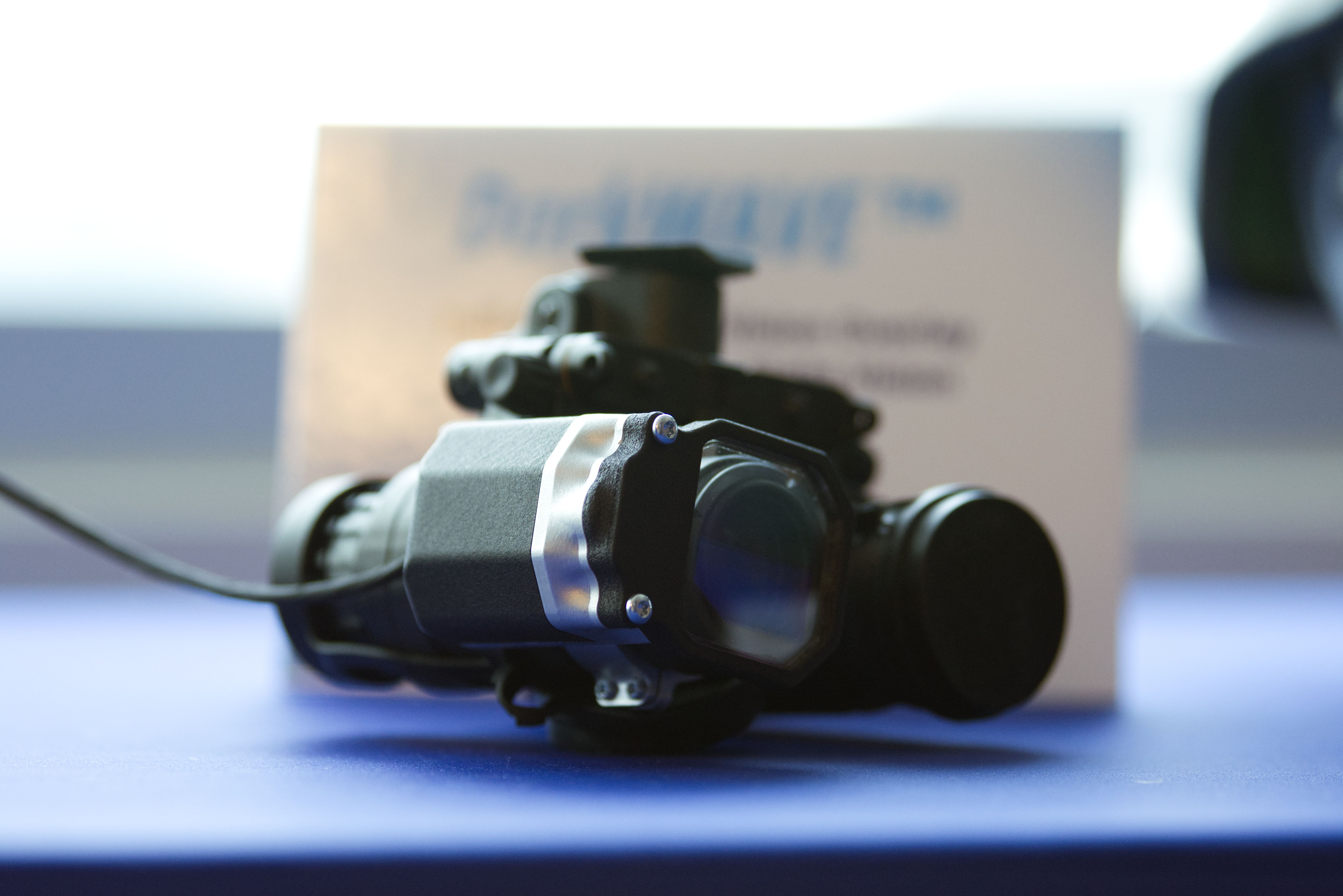 Night-vision goggles with a clip-on enhancement sit on a blue table. The background is washed out with light.