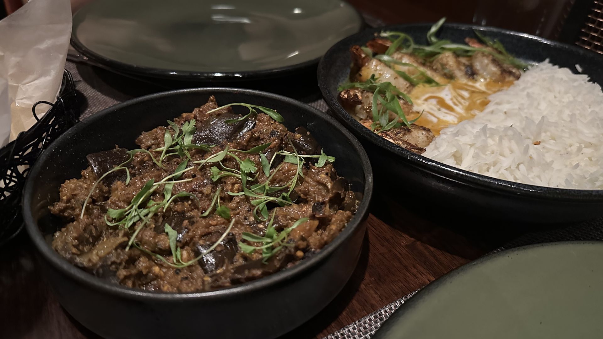 A bowl of eggplant Hyderabad is seen on a wooden table, with dinner plates next to it. In another bowl is a shrimp dish with rice.