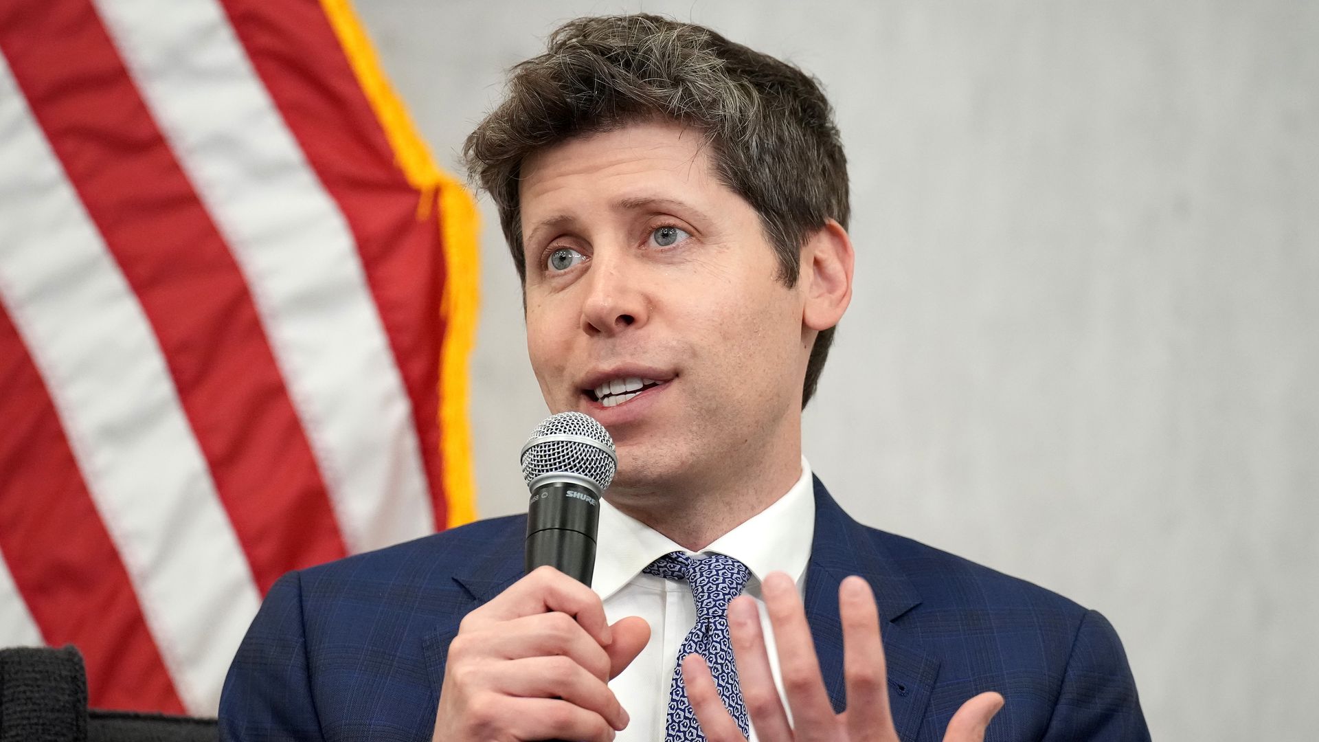 OpenAI CEO Sam Altman, in a suit and holding a microphone, seated in front of an American flag. 