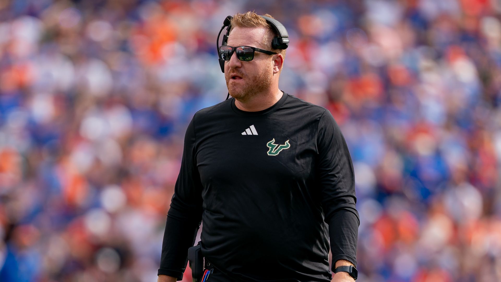University of South Florida Head football coach Alex Golesh stands in a football stadium, with a blurred crowd in the background. 