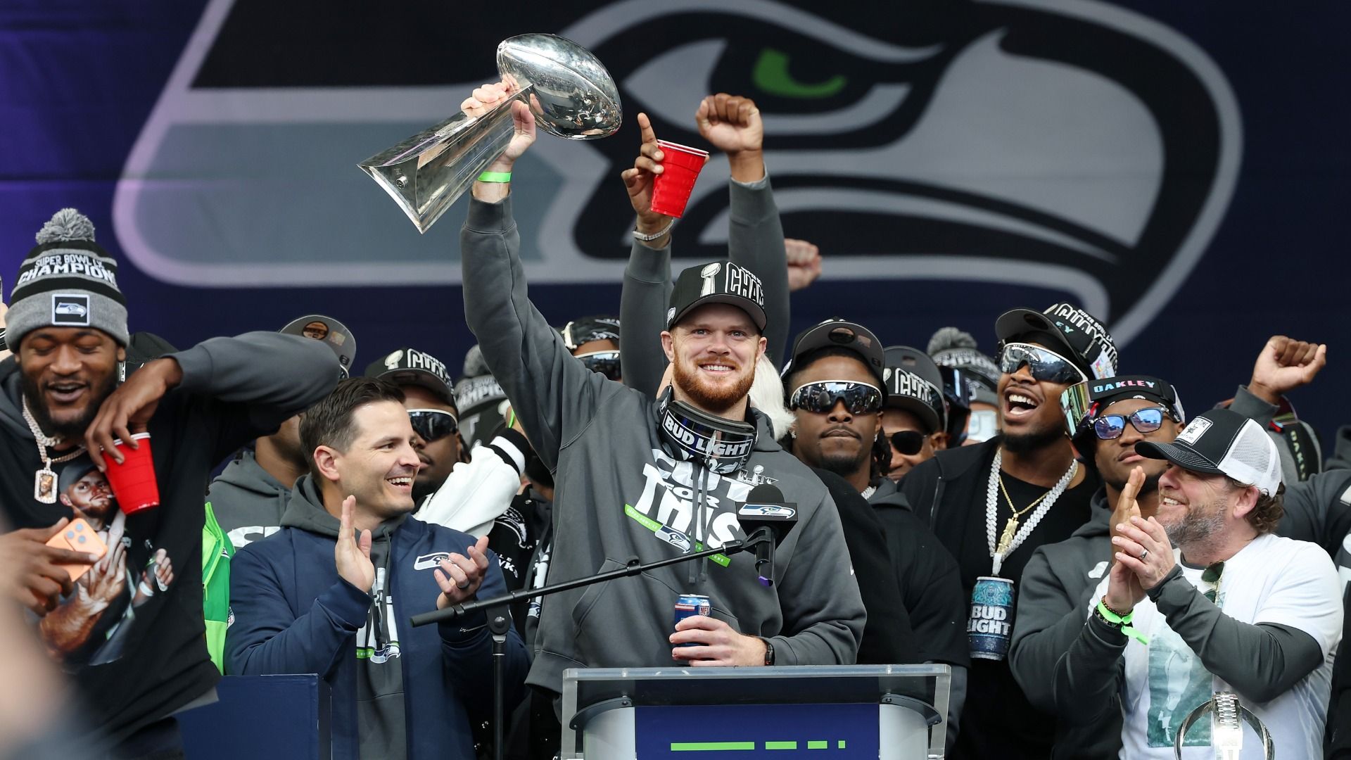 Sam Darnold wears a gray sweatshirt while holding up the Lombardi Trophy at a podium. He is surrounded by his teammates, with a Seahawks logo behind him. 