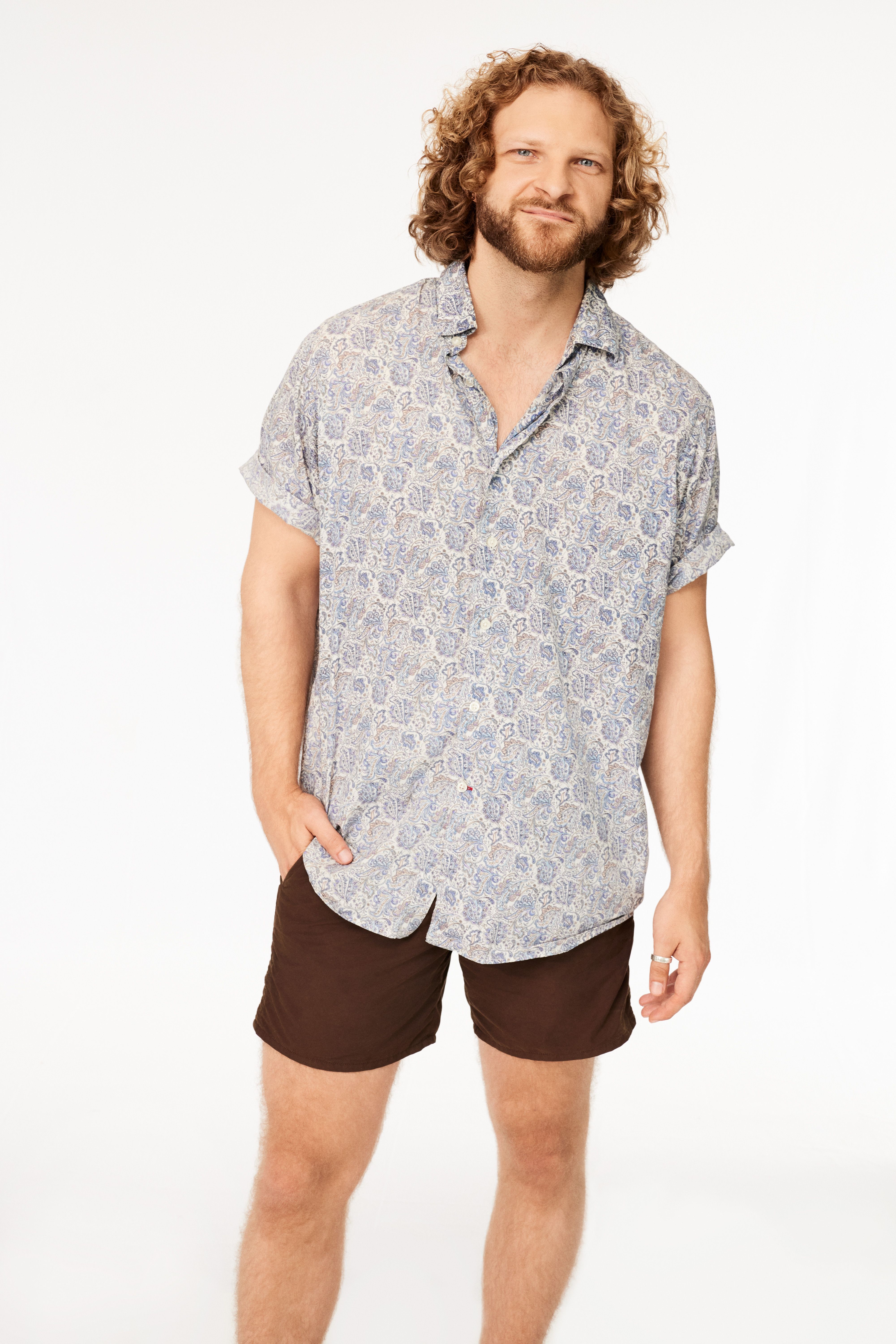 Man with curly hair and beard wearing a rolled-sleeve blue patterned shirt and brown shorts, standing against a white background with one hand in his pocket.