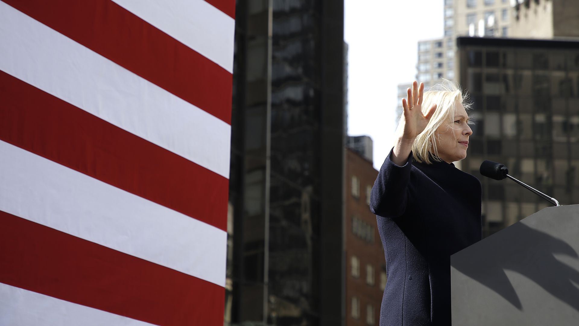 Sen. Gillibrand stands and waves in front of a podium with a microphone, with an American flag behind her.