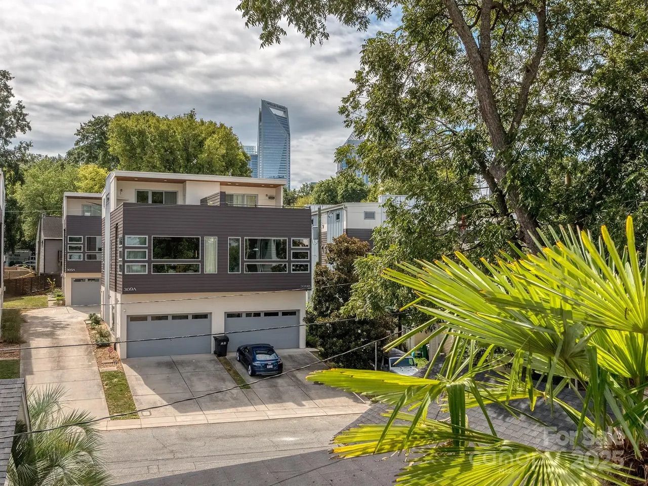 Modern two-story gray and white duplex with large windows and garages, surrounded by green trees, palm plants in foreground, under cloudy sky near a city skyline.
