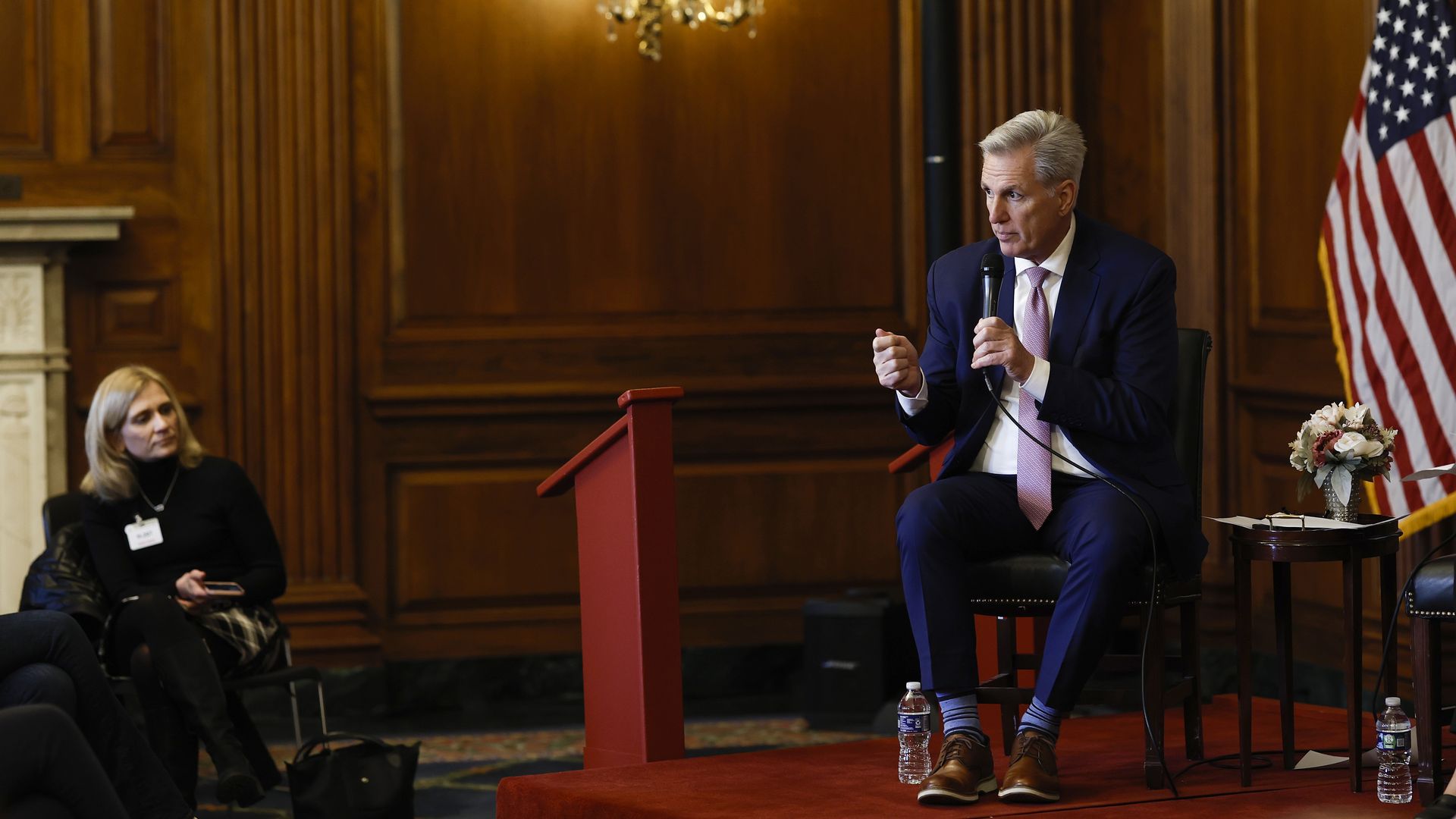 House Speaker Kevin McCarthy, wearing a blue suit, white shirt and pink tie, speaks into a microphone while sitting in a chair next to an American flag.