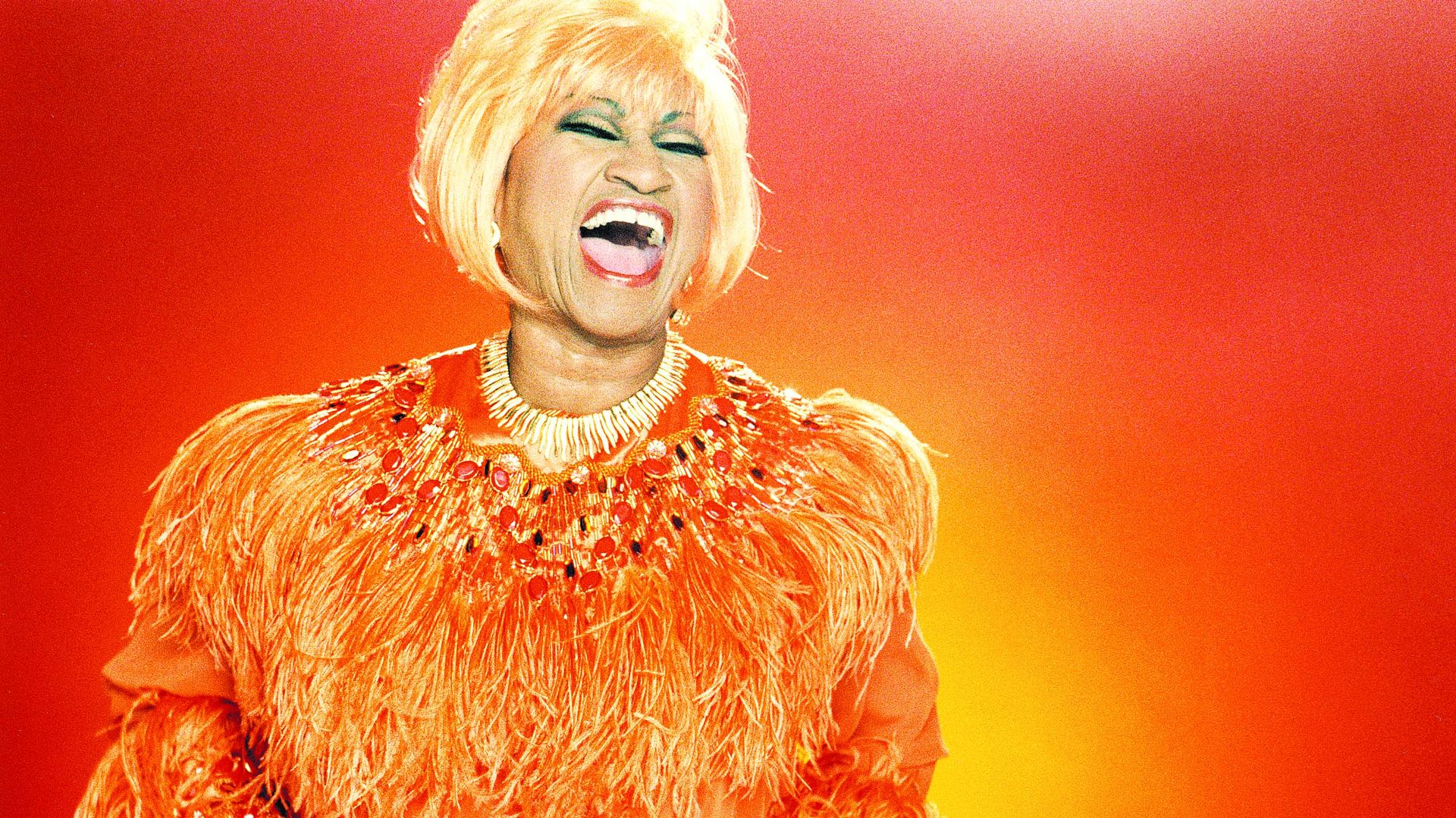 Celia Cruz wears a blonde bob wig and an orange dress. 