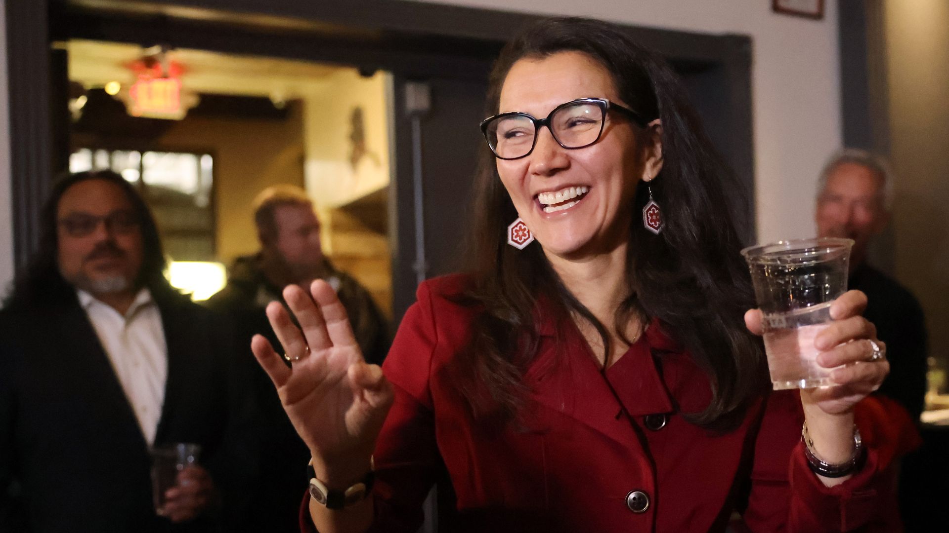 Smiling woman in a red coat holding a clear plastic cup with water, wearing glasses and red patterned earrings, waving with her other hand indoors with blurred people in the background.